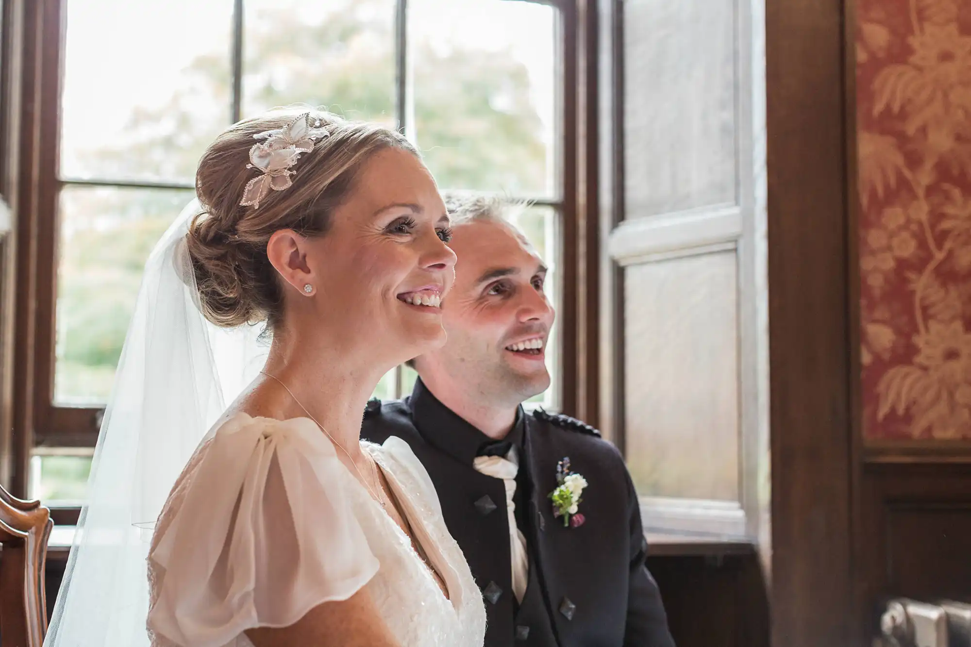 A bride and groom smiling and looking out a window, the bride in a veil and the groom in a black suit.