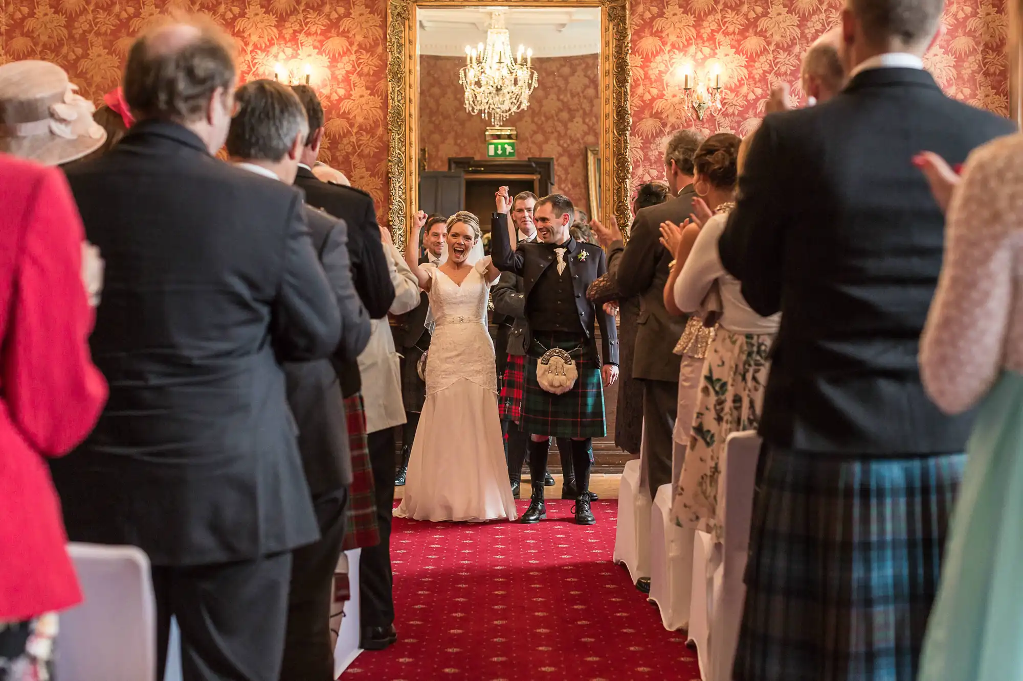 A newlywed couple walks down the aisle, smiling, as guests applaud. the groom is in a kilt and the bride in a white dress.