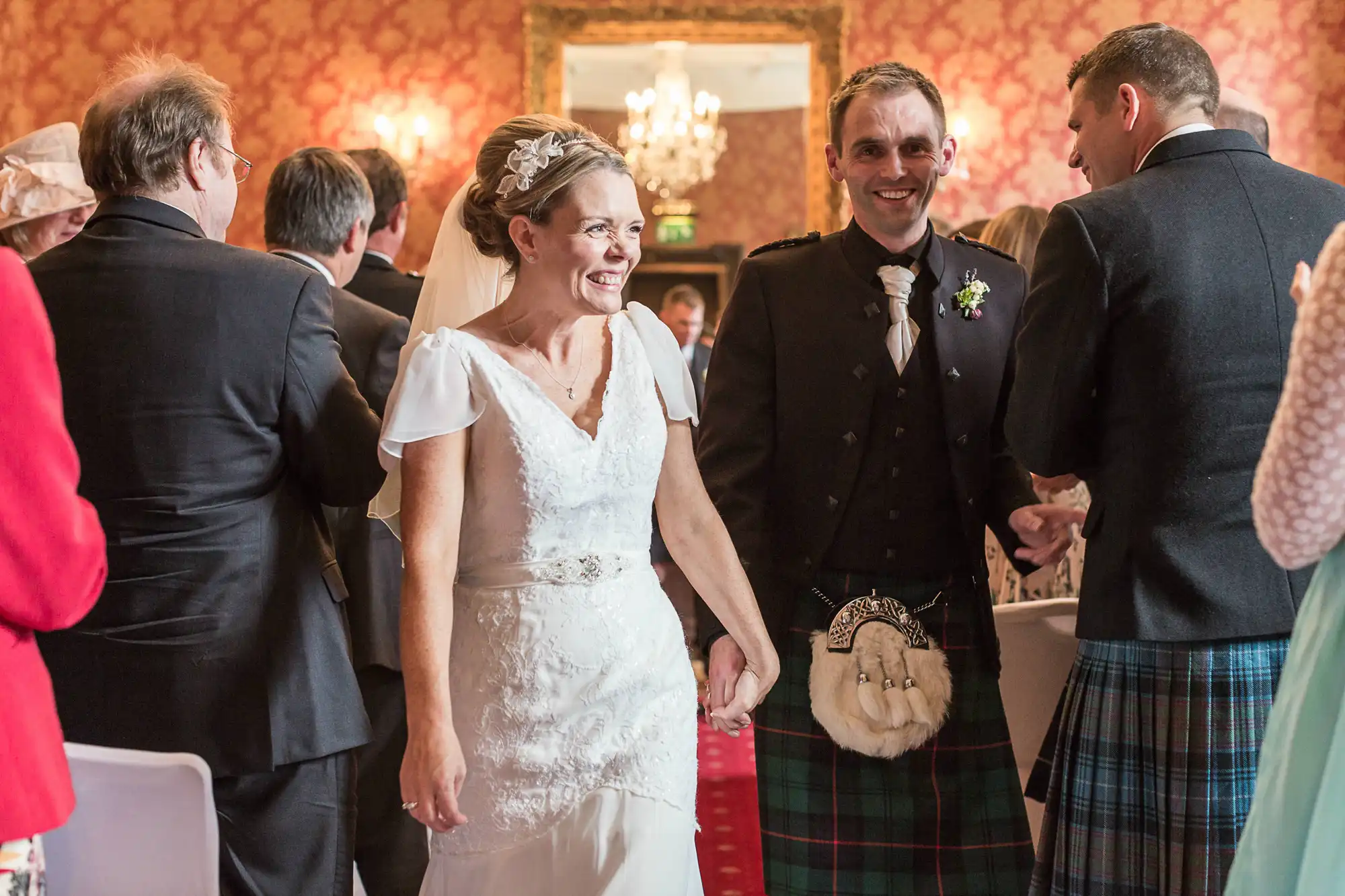 A bride in a white dress and a groom in a scottish kilt, both smiling, walk hand in hand through a crowd at a wedding reception.
