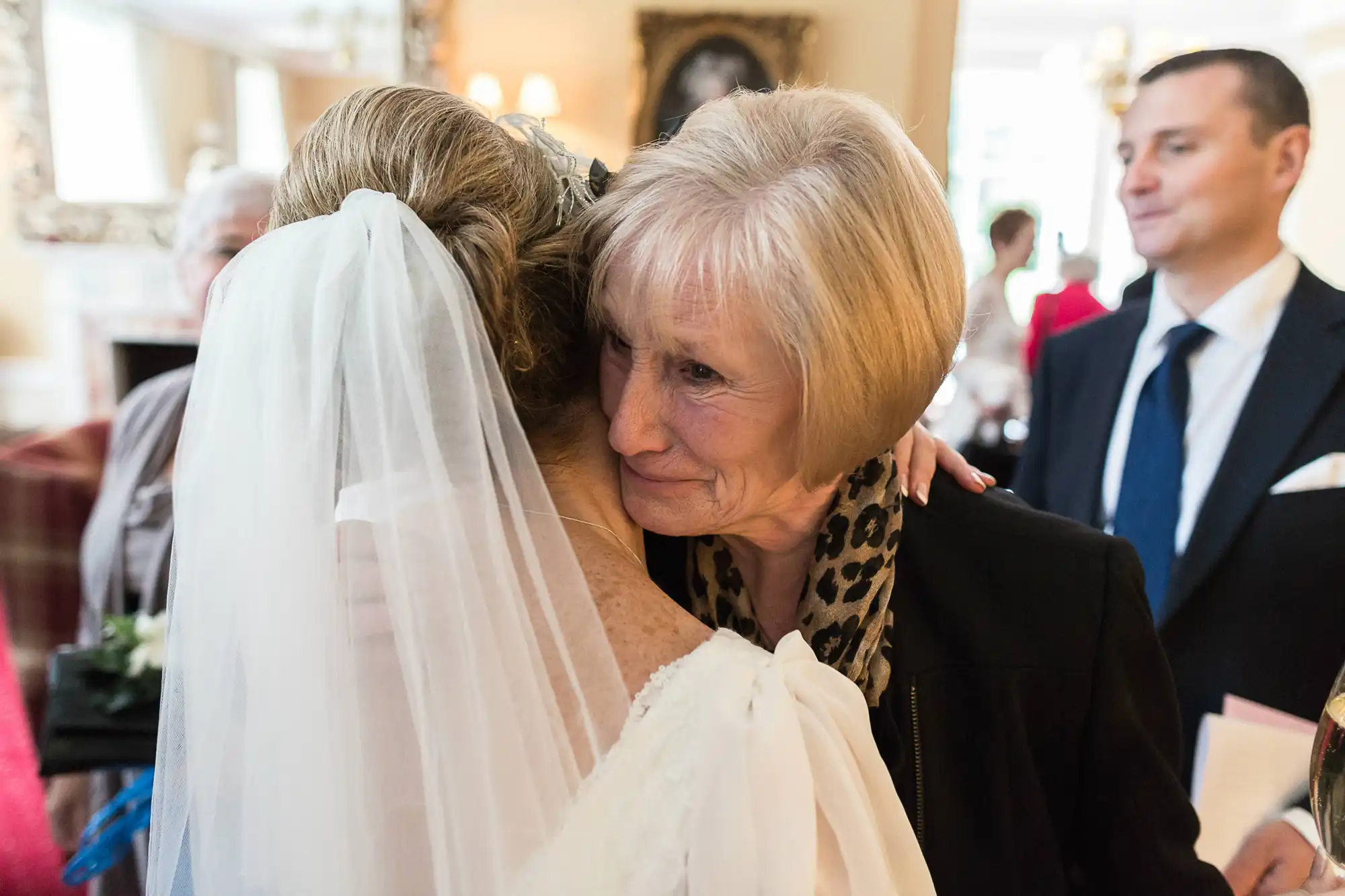 An older woman with a tender expression hugs a bride in a white dress and veil, surrounded by guests at a wedding reception.