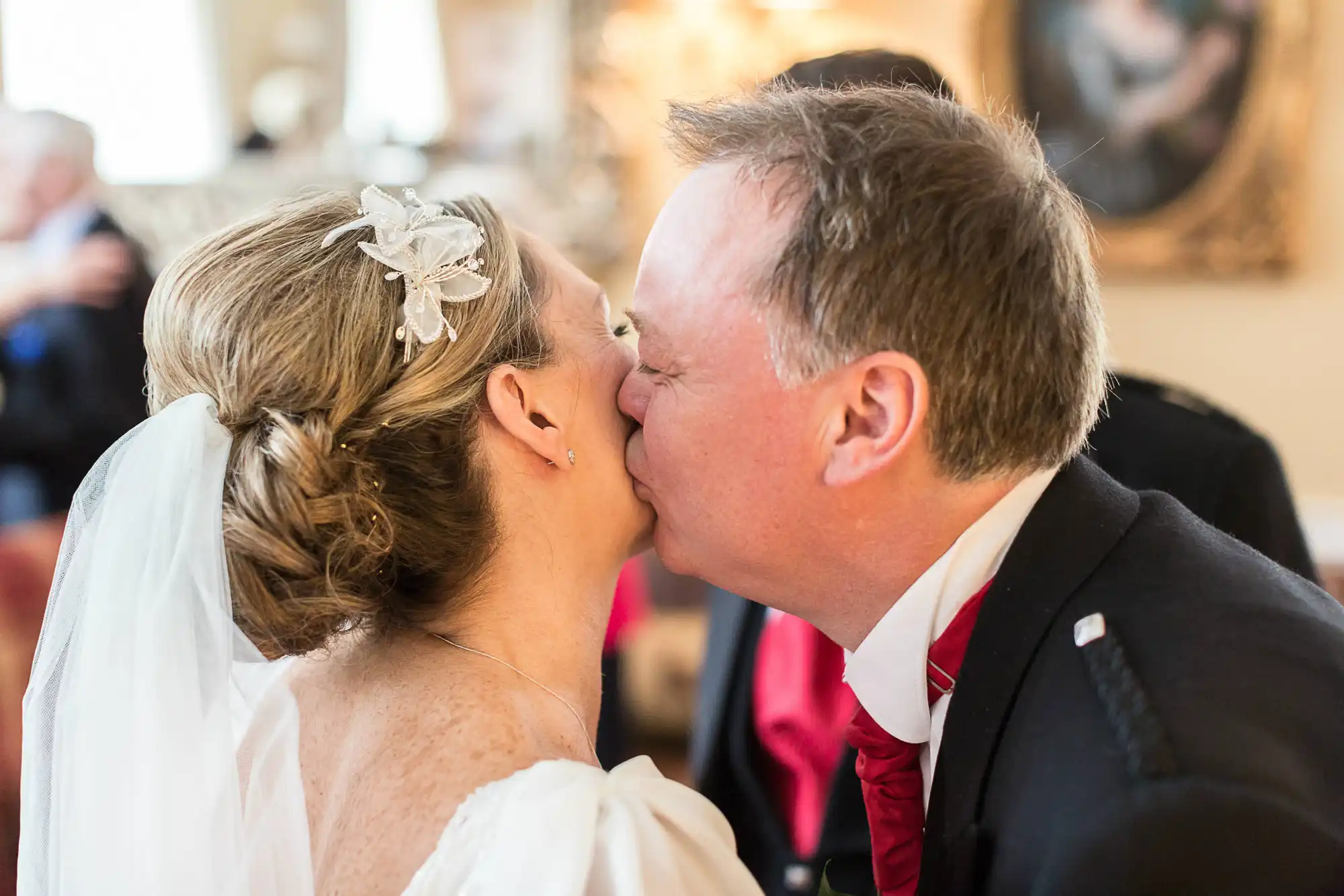 A bride and groom kiss tenderly on their wedding day, surrounded by a blurred background of guests.