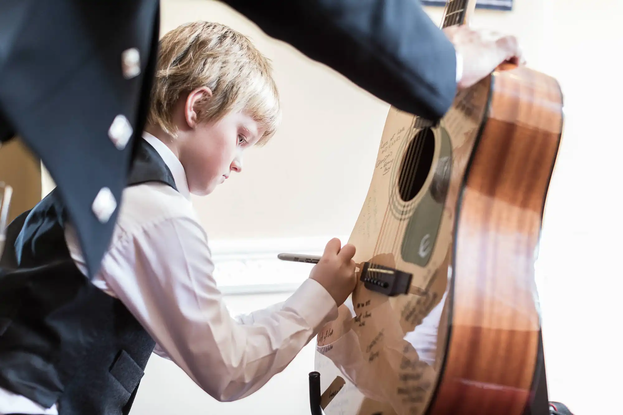 A young boy in a suit signs an acoustic guitar while looking intently at where he writes.