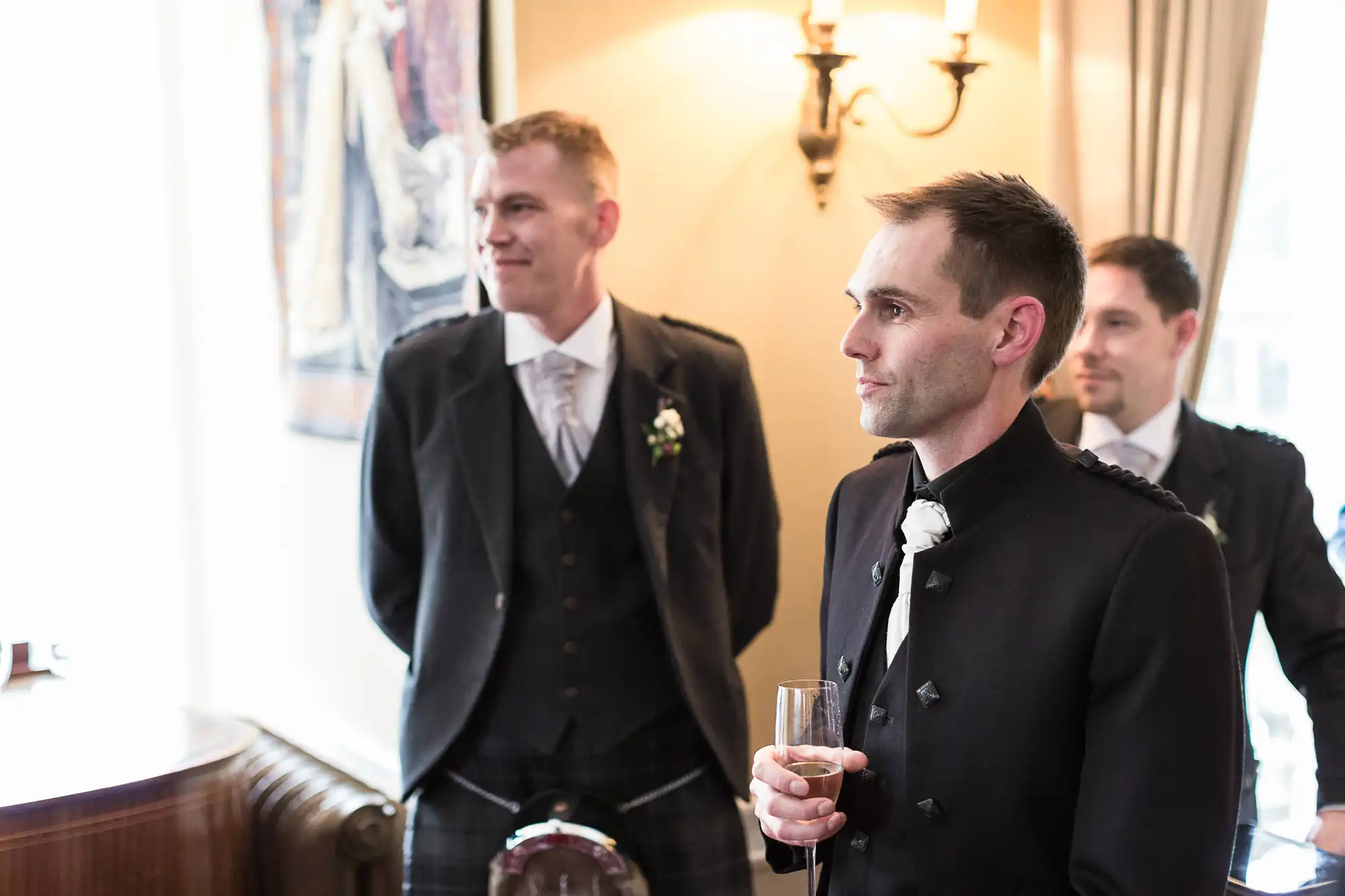 Three men in formal attire at an indoor event, with the nearest holding a champagne flute.