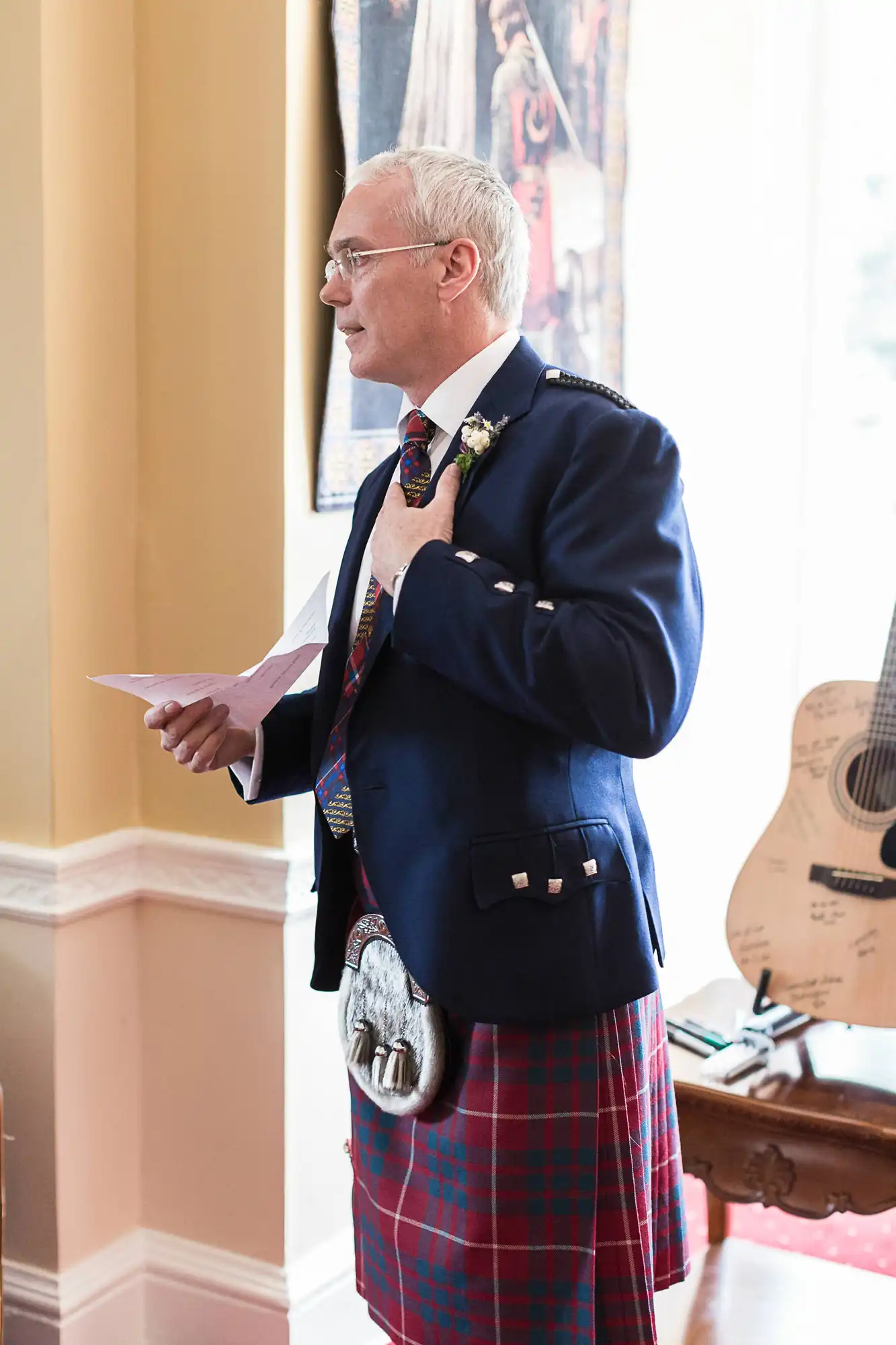 A man in a traditional scottish outfit, featuring a kilt and sporran, stands in a room holding a speech note, with a guitar in the background.