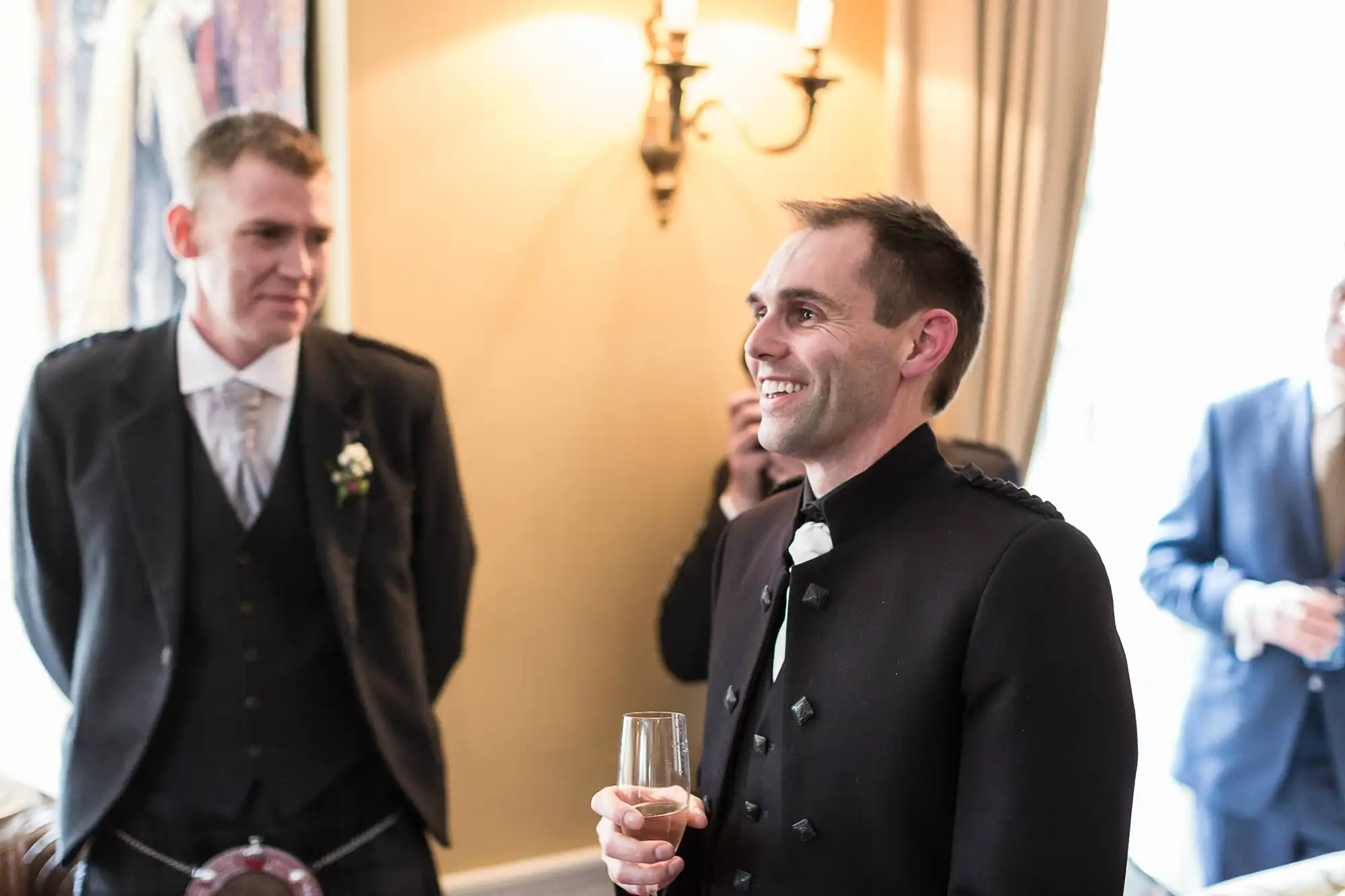Two men in formal attire at an indoor event, one holding a champagne glass and smiling, with elegant lighting in the background.