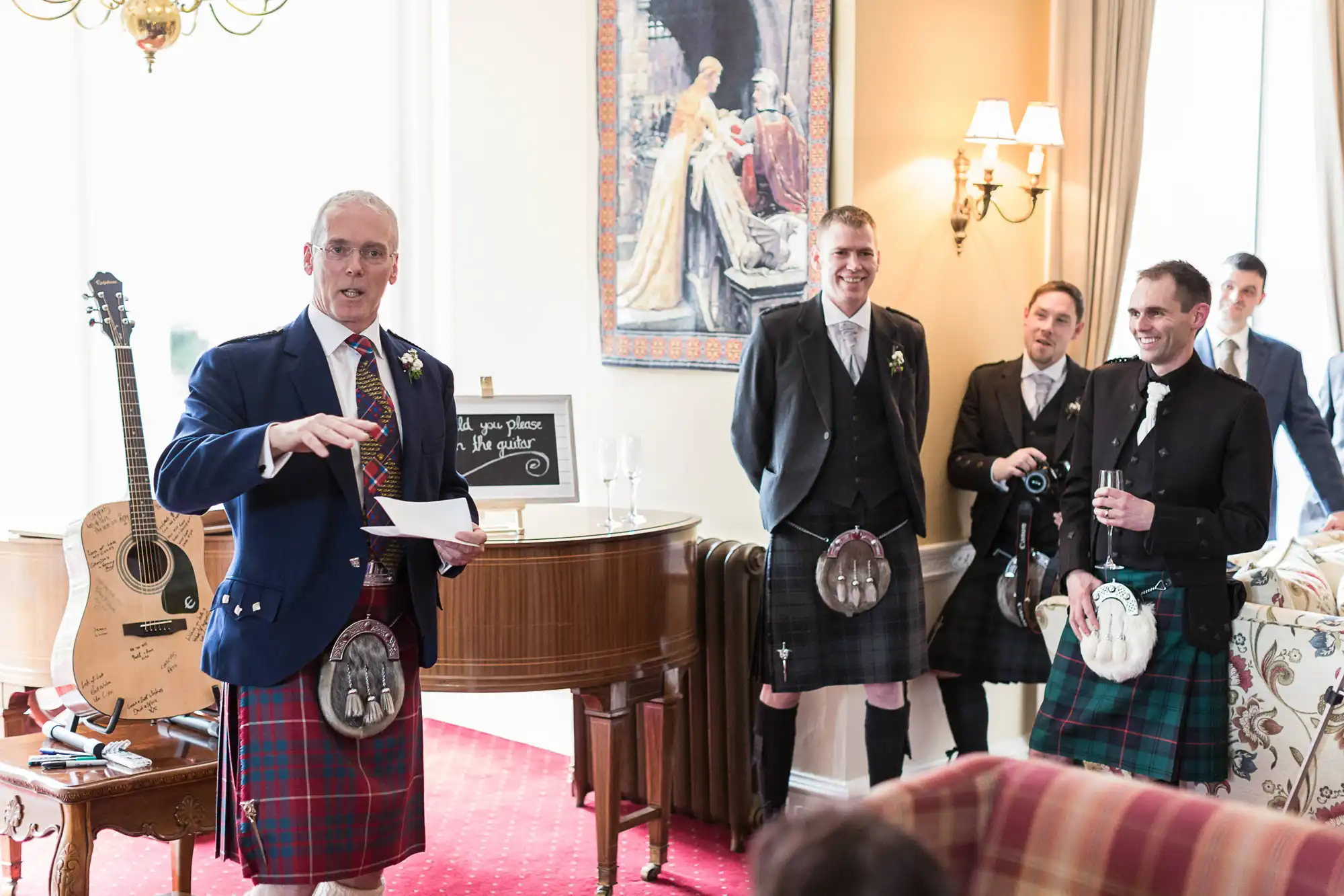 A man in a kilt speaking at a wedding event, holding a piece of paper, with three other men in kilts smiling around him in a decorated room.