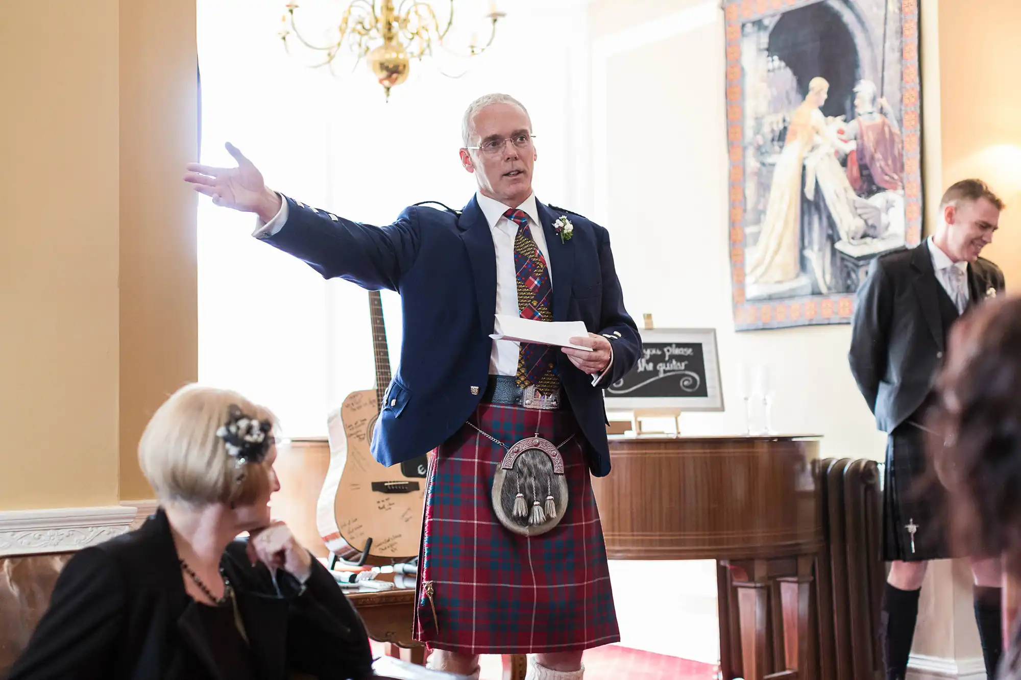 A man in a kilt and formal jacket gestures while speaking in front of a group of seated people in an elegant room.