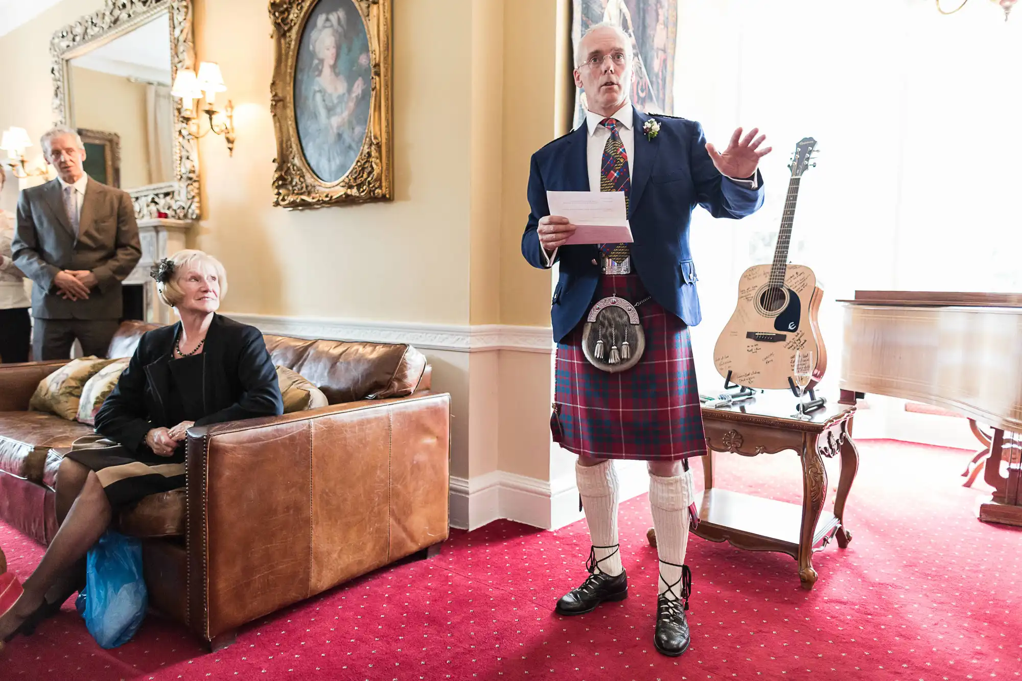 An elderly man in a kilt and sporran speaks from a paper in a formal room with seated listeners, portraits on the wall, and a guitar on display.