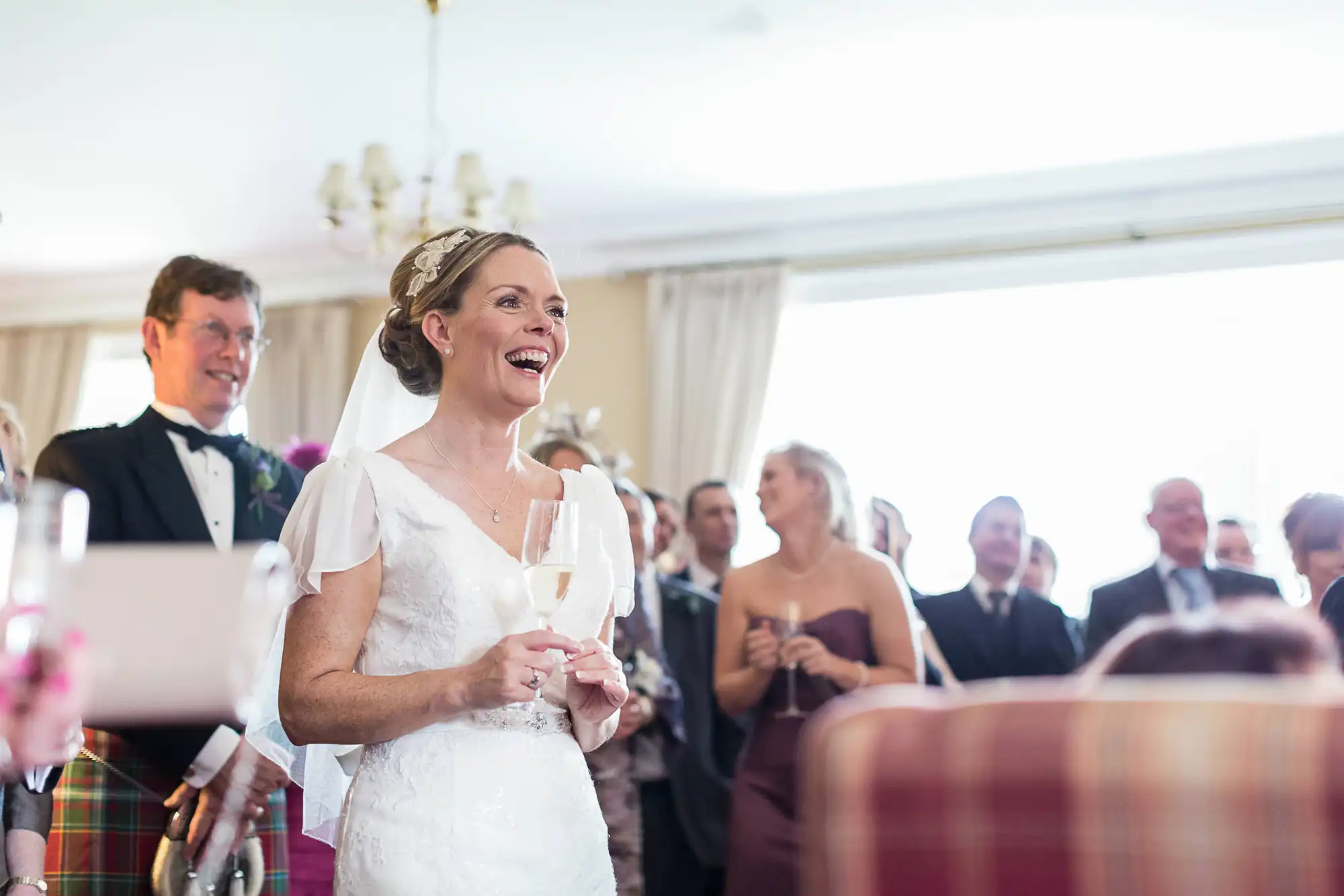 A bride in a white dress smiling joyfully while holding a champagne glass, with guests and a man in a kilt in the background at a wedding reception.