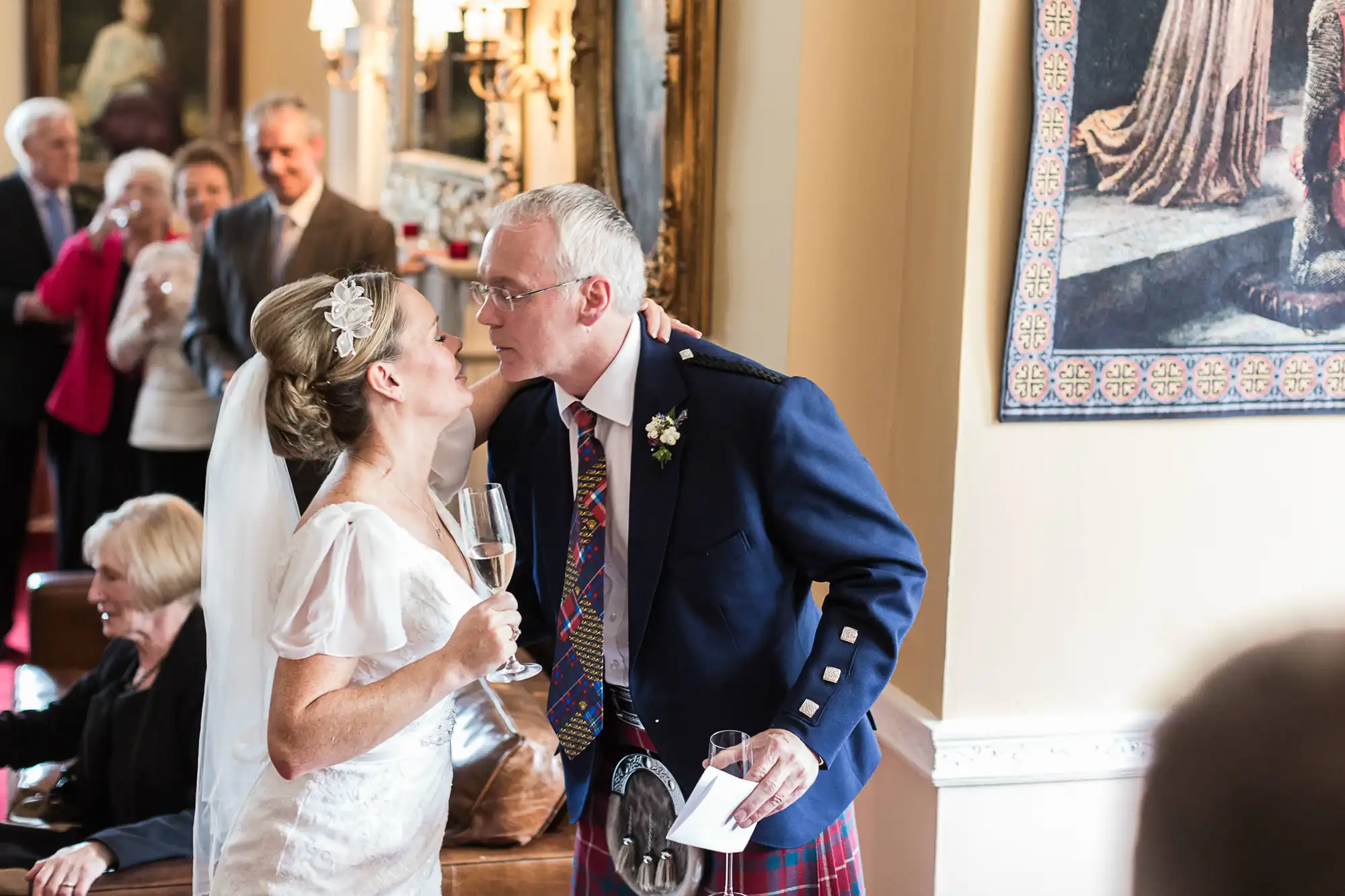 A bride and groom, dressed in a white gown and scottish kilt, respectively, sharing a kiss while holding wine glasses at their wedding reception.