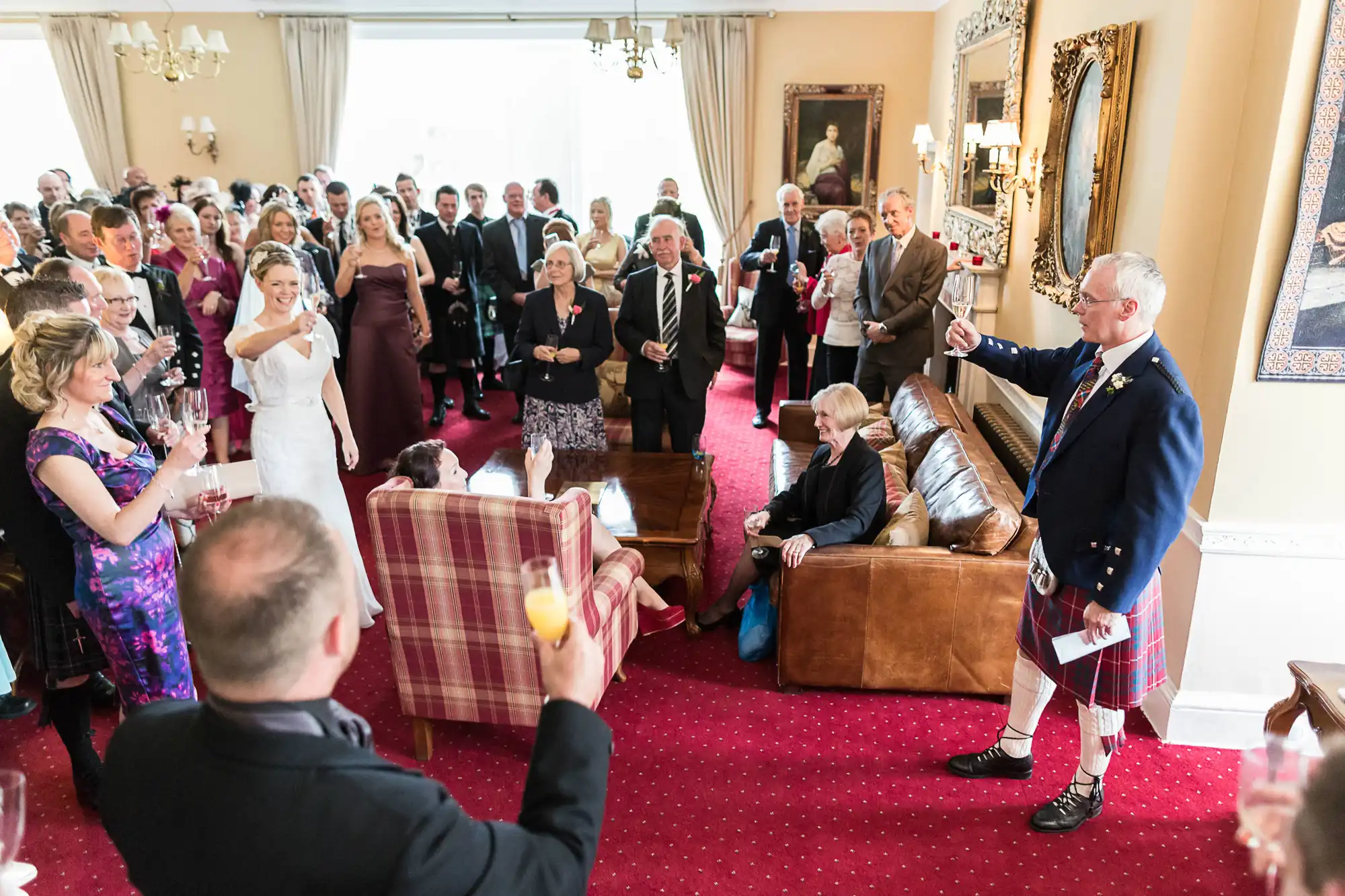 A wedding reception in an elegant room with guests watching a man in a kilt giving a speech.