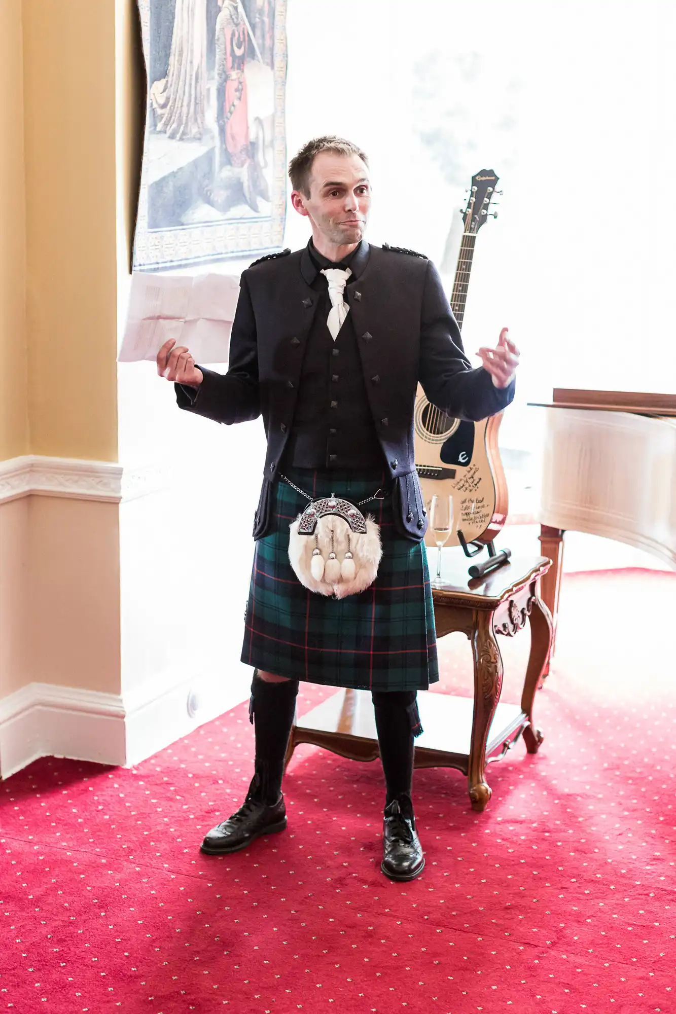Man in traditional scottish attire, including kilt and sporran, standing in an elegant room with a guitar on a stand behind him.