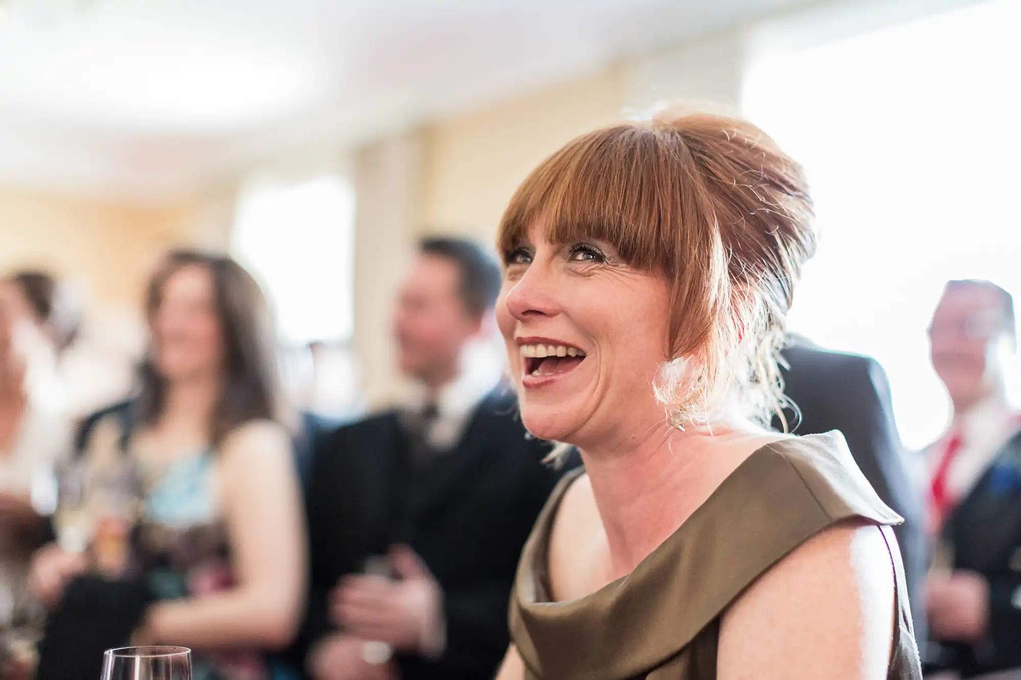 Woman with short red hair laughing at a social event, wearing an elegant brown dress. blurred guests in the background.