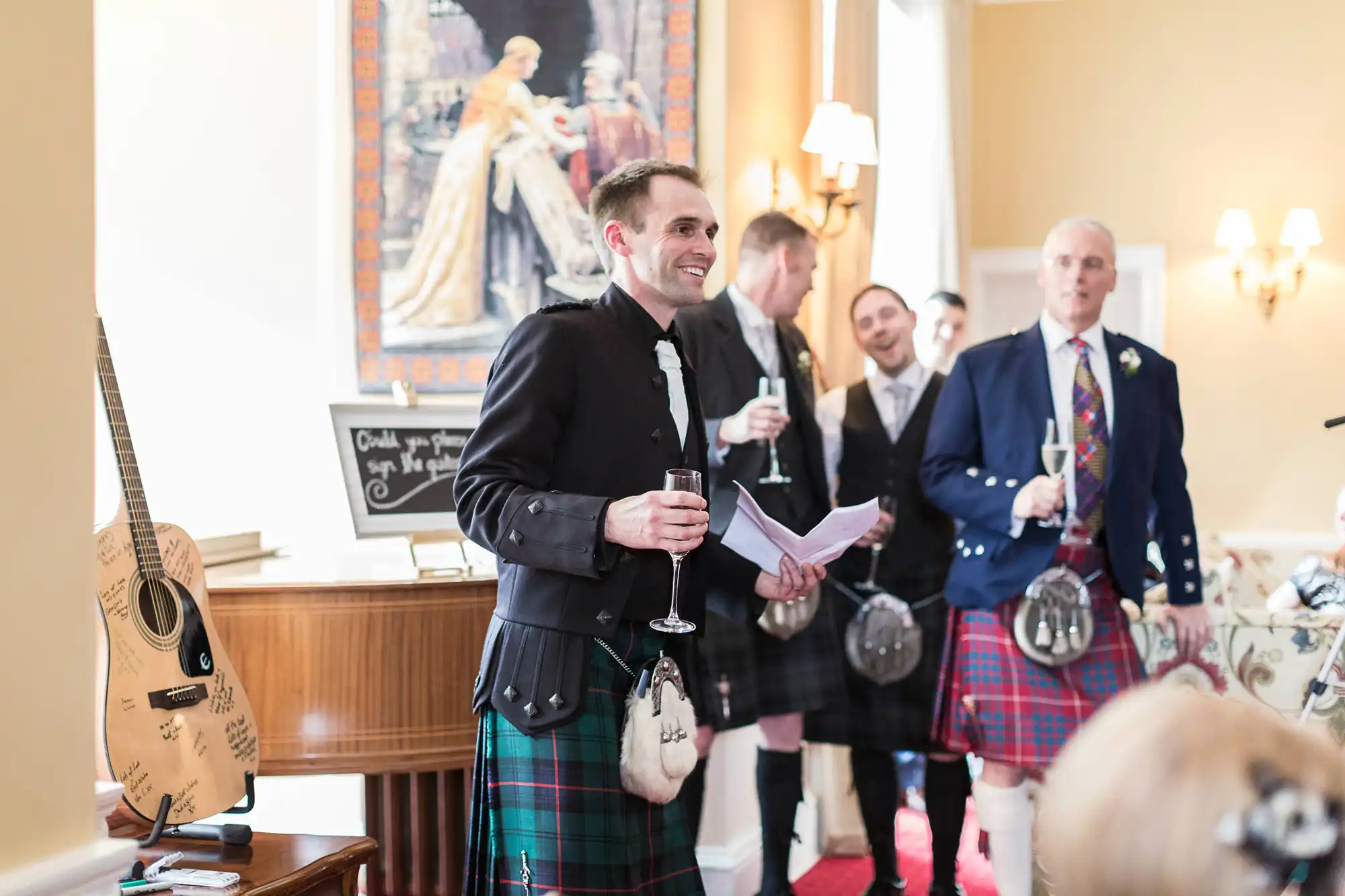 Man in kilt holding a speech and a glass of wine, smiling at a wedding reception with other guests in kilts around him.