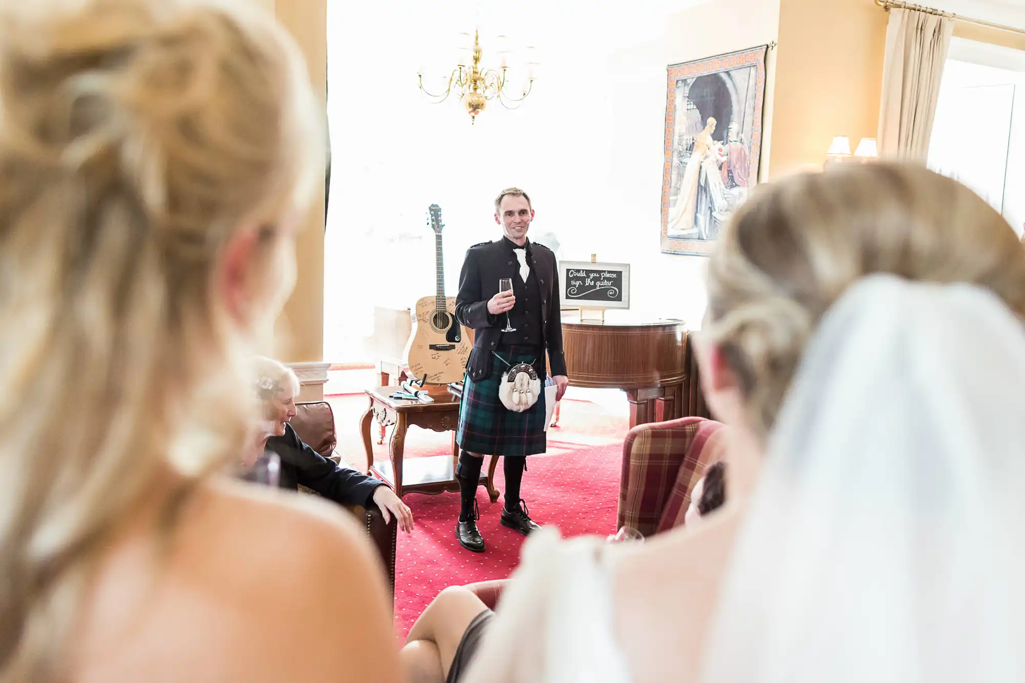 A groom in a kilt gives a speech to a bride and guests in an elegant room with a chandelier and classical guitars in the background.