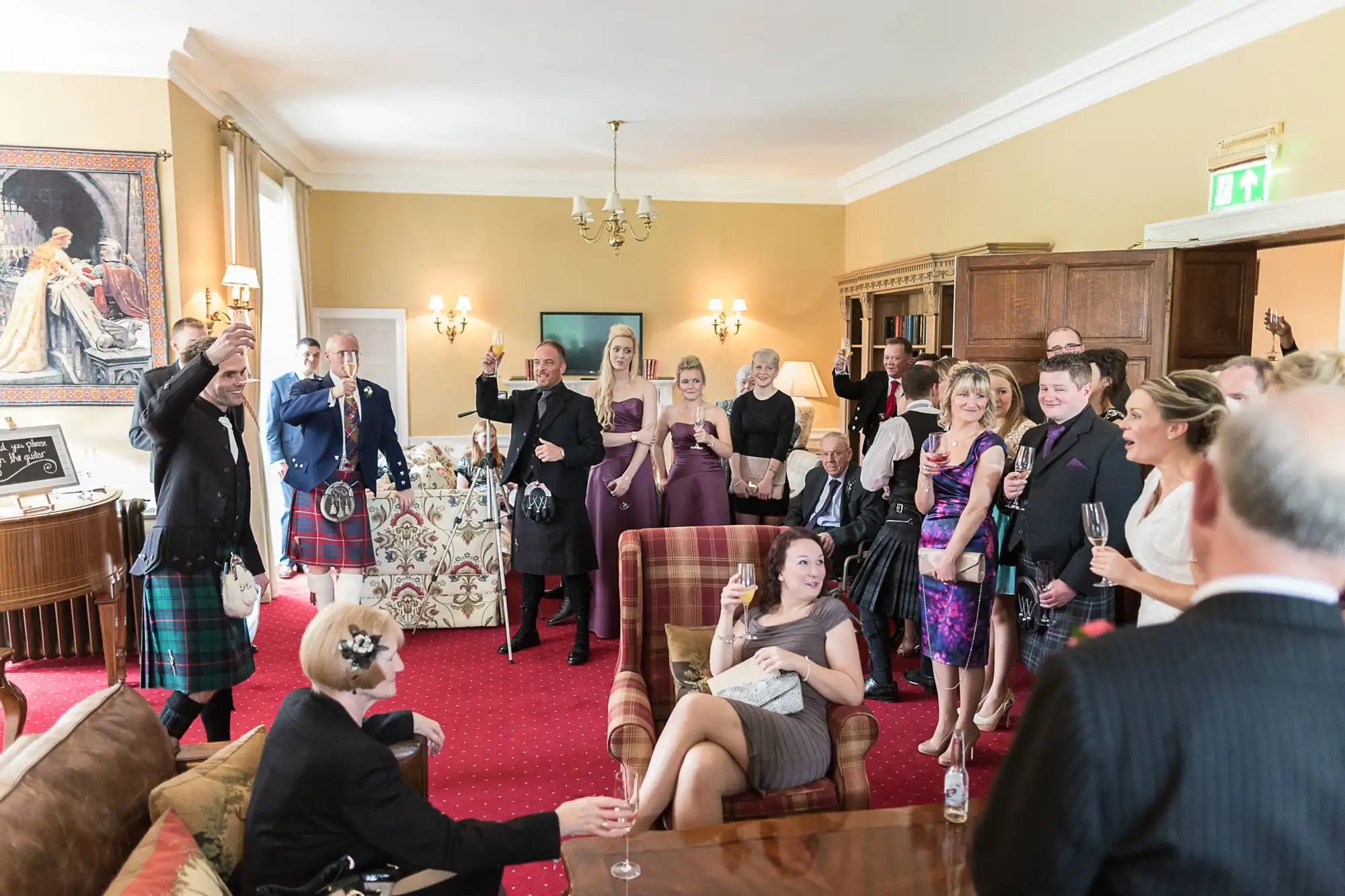 A group of elegantly dressed people holding drinks and enjoying a lively gathering in an opulent room, some standing, one sitting on a couch, with a few wearing traditional kilts.