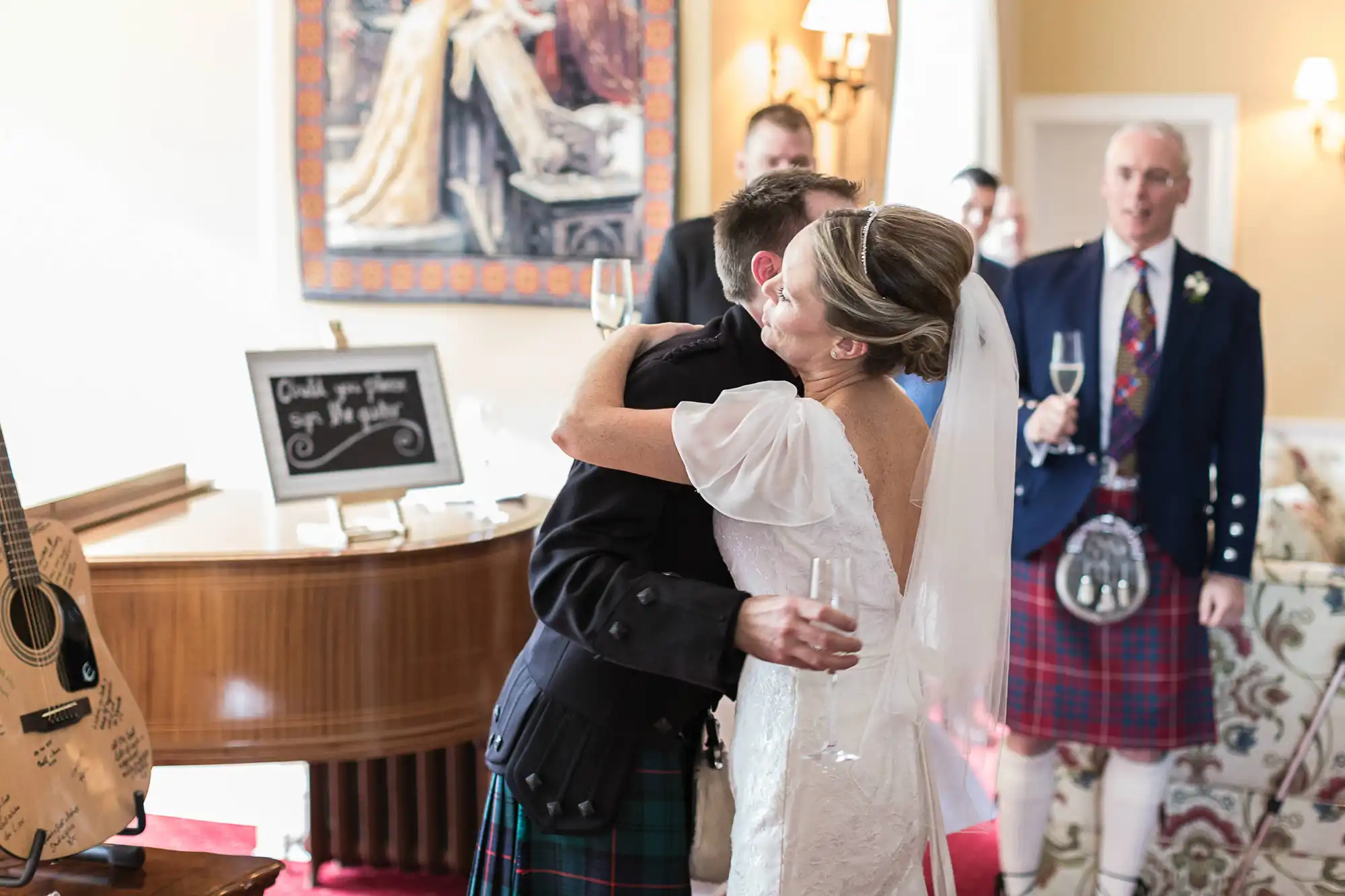 A bride in a white dress and a groom in a kilt embrace warmly, surrounded by wedding guests in a decorated room.