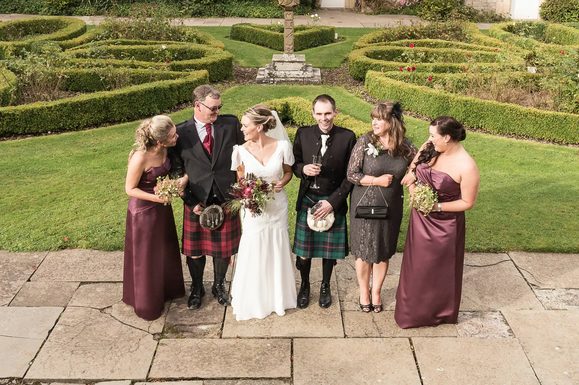 A wedding group photo in a garden, featuring a bride and groom with four guests, two men in kilts and three women in burgundy dresses, holding bouquets.