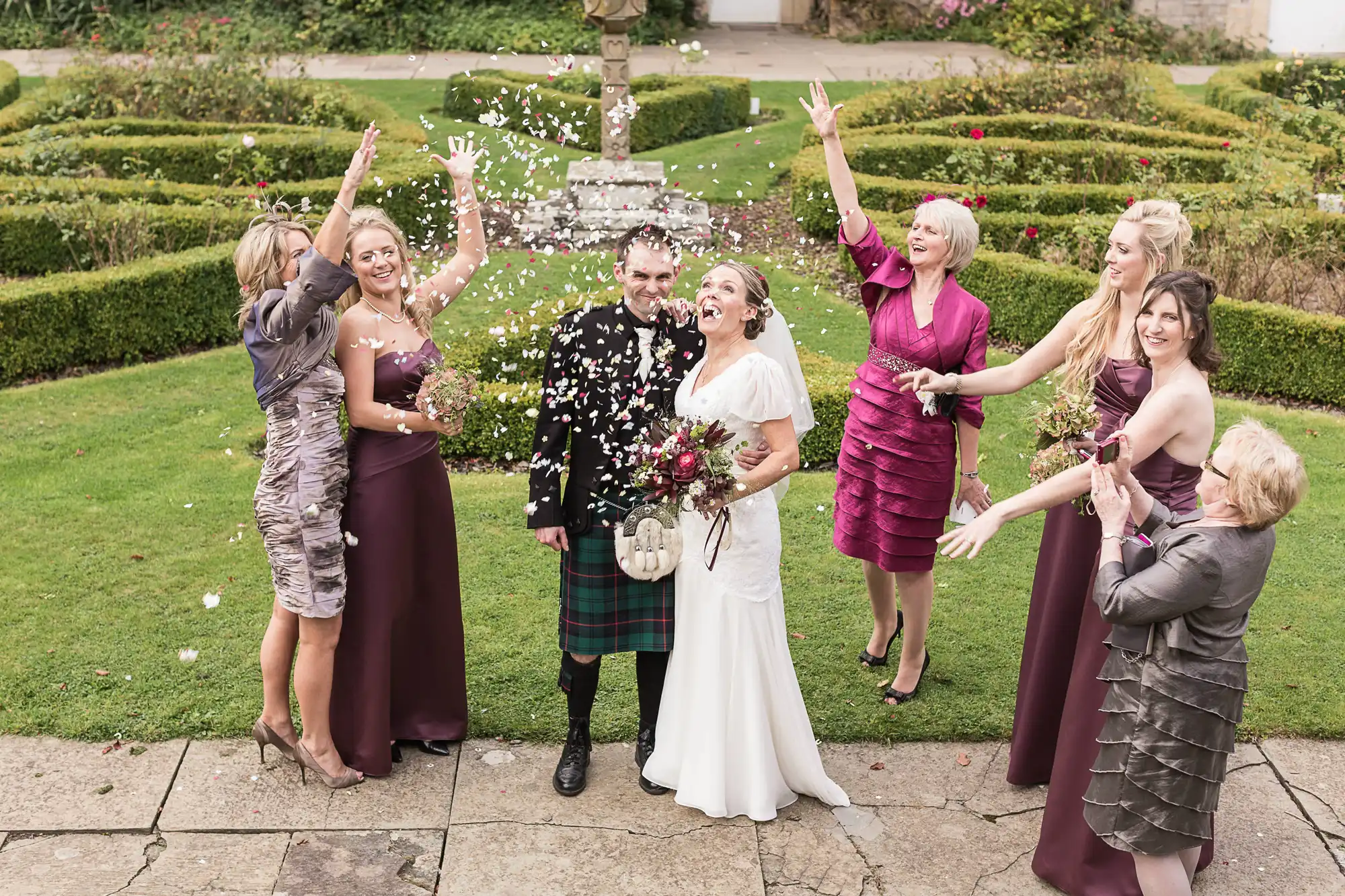 A joyful wedding group celebrates by throwing confetti at the smiling bride and groom in a garden setting.