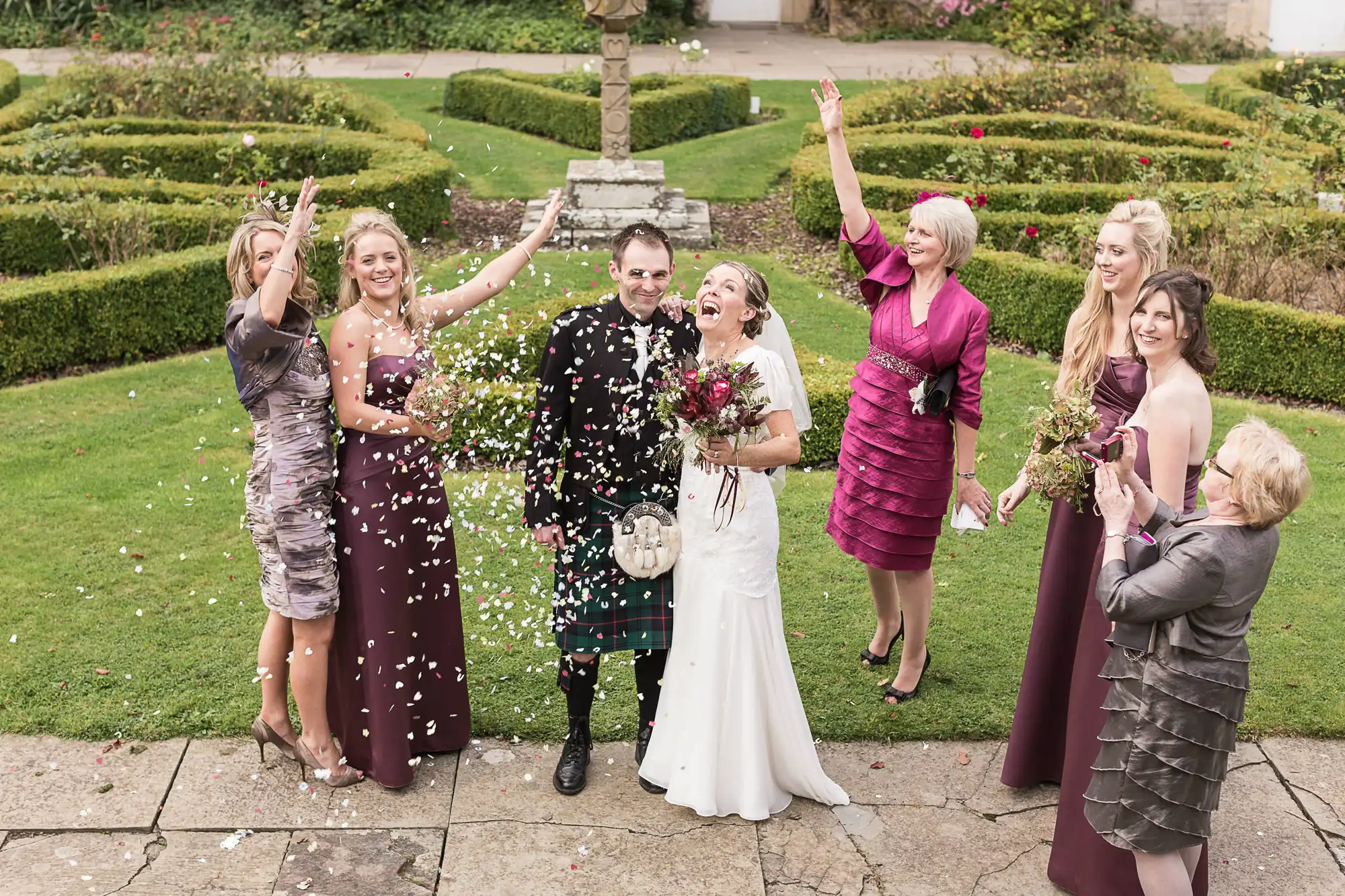 A wedding party joyfully throwing flower petals at a newlywed couple in a garden, with guests smiling and celebrating.