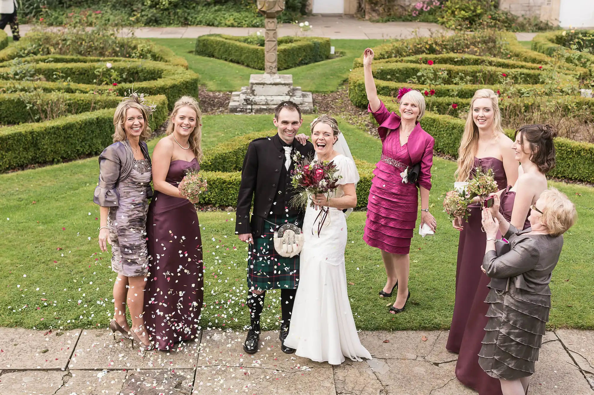 A joyful wedding scene with a bride and groom smiling among a group of five women throwing flower petals in an ornate garden.