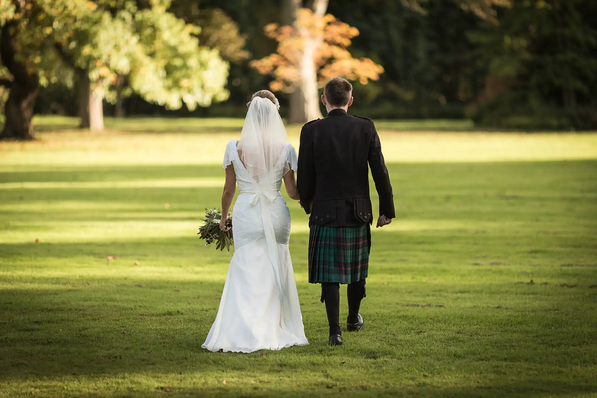 A bride in a white gown and a groom in a kilt and jacket walk hand in hand across a sunlit, grassy park.