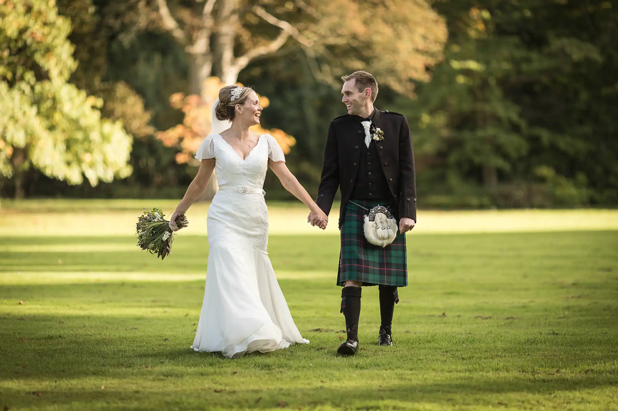 Bride in a white dress and groom in a kilt holding hands and smiling while walking through a park.