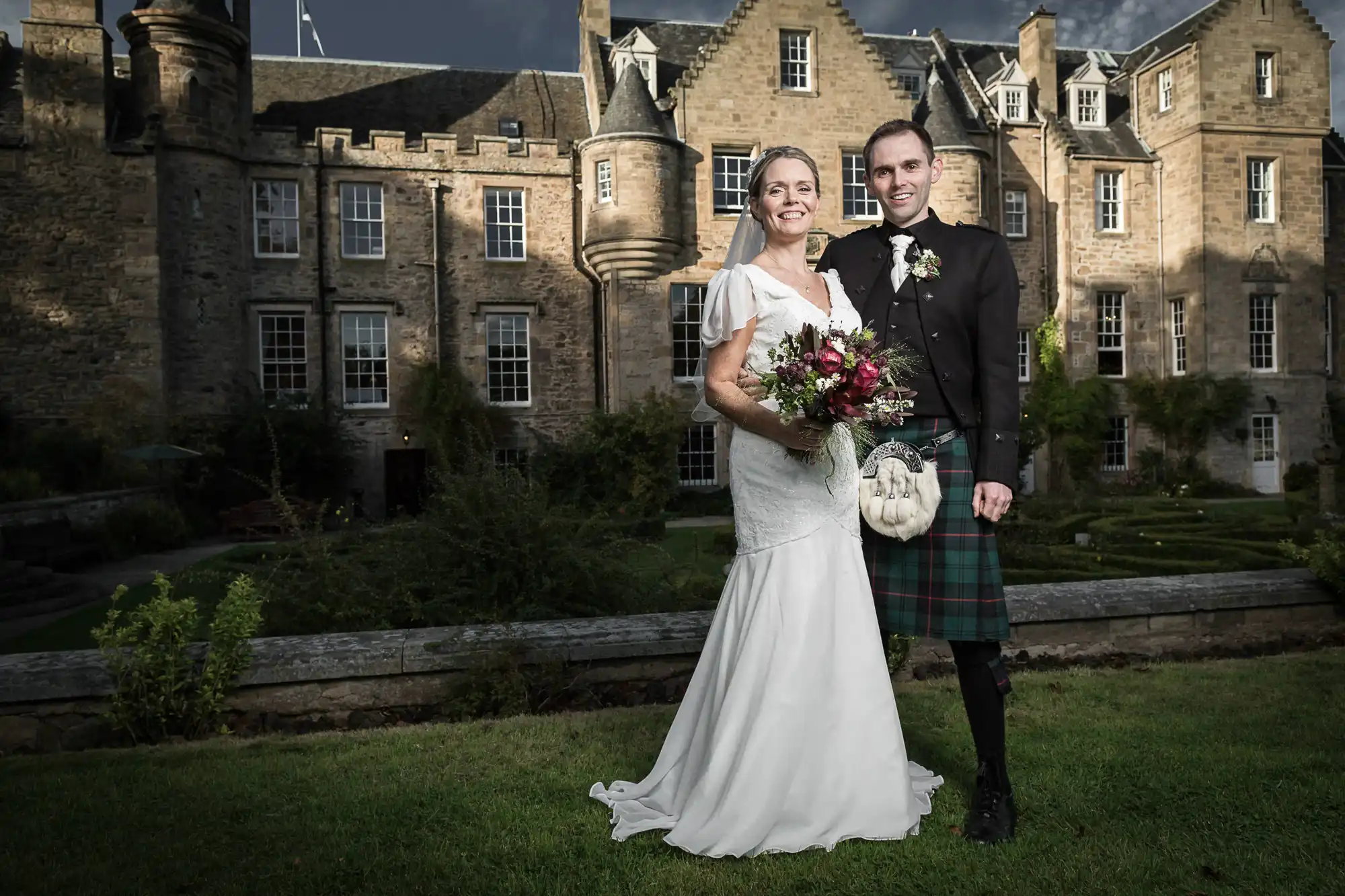 A bride and groom in wedding attire, the groom wearing a kilt, stand smiling in front of a historic stone castle under a cloudy sky.