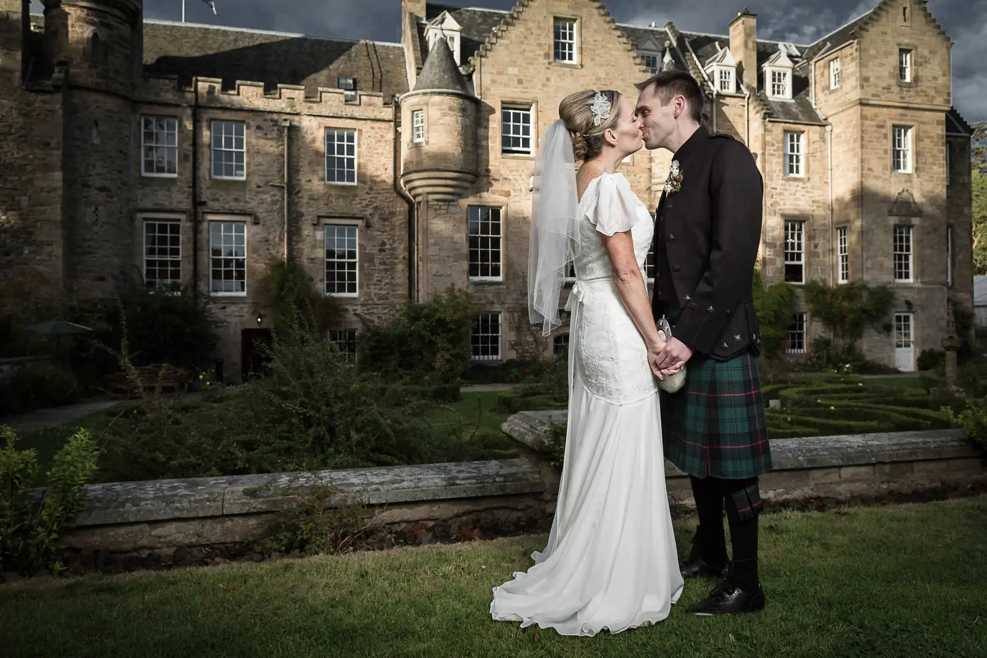 A bride in a white dress and a groom in a kilt kissing in front of an old castle with manicured gardens under a cloudy sky.