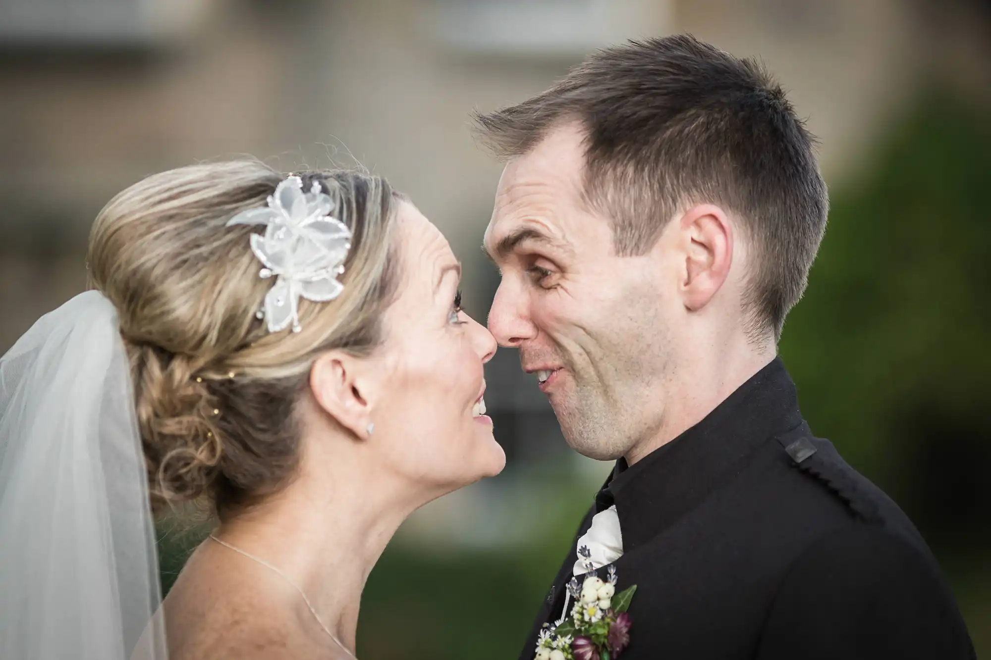 A bride and groom lovingly touch noses and smile at each other, dressed in wedding attire, with a soft-focus background.