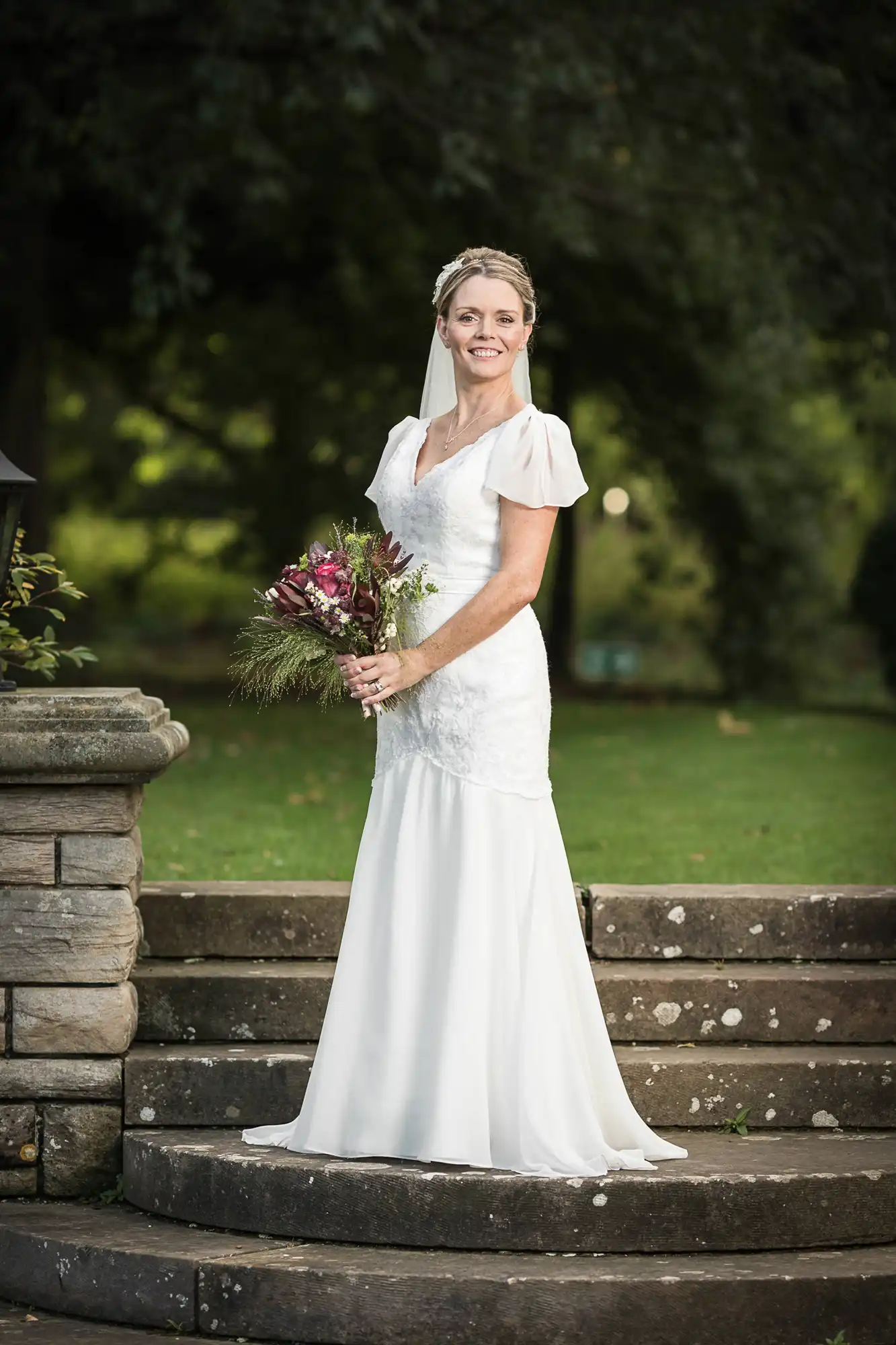 A bride in a white wedding dress holding a bouquet stands on stone steps in a park.