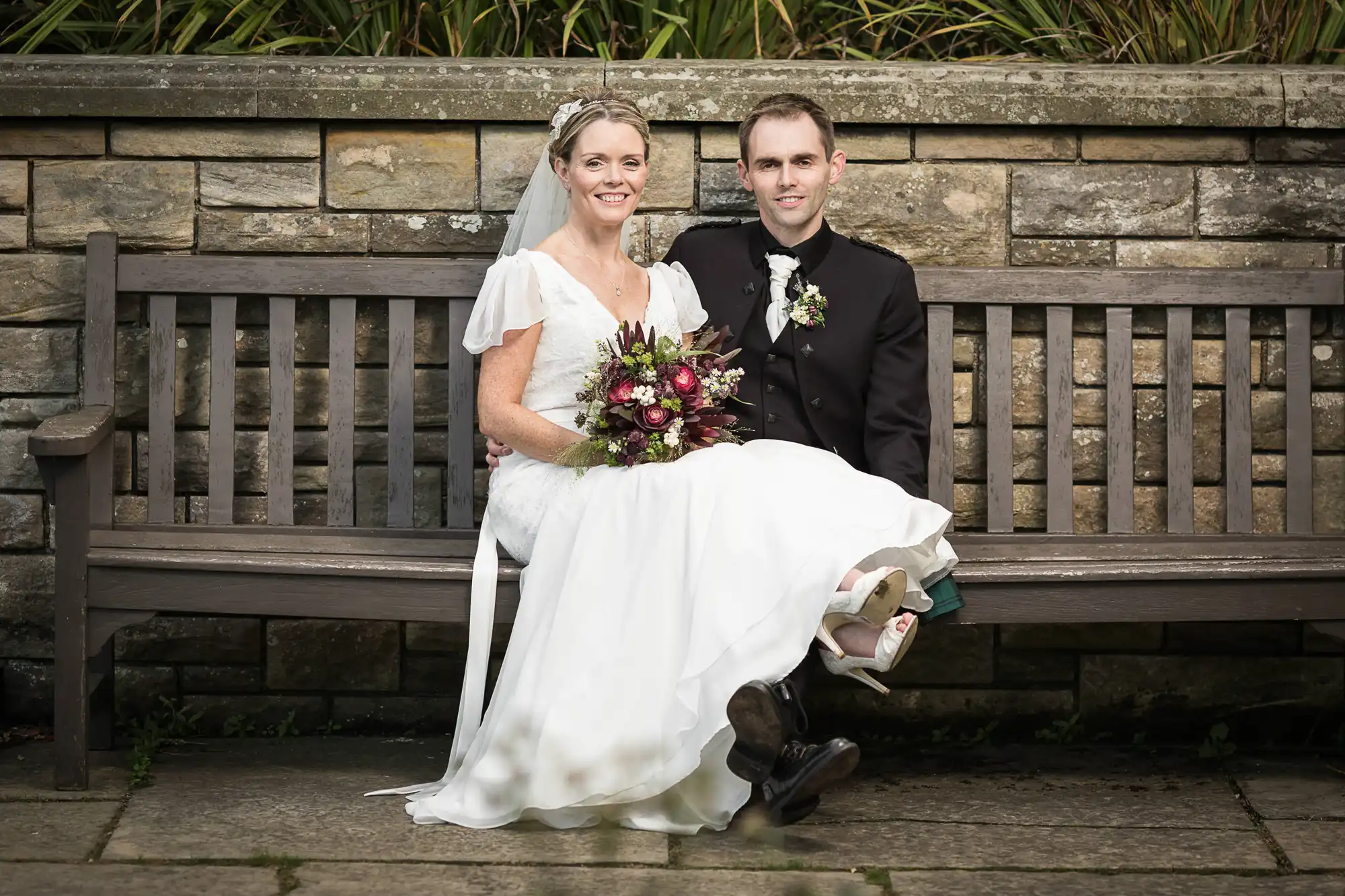 A bride and groom sitting on a bench, the bride in a white gown holding a bouquet and the groom in a dark suit, both smiling.