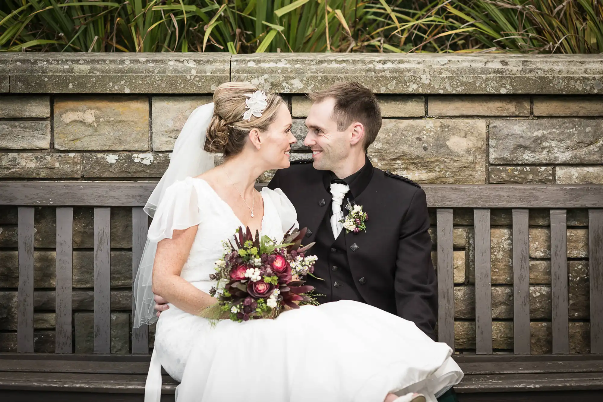A bride and groom smiling at each other while sitting on a bench, the bride holding a bouquet of flowers.