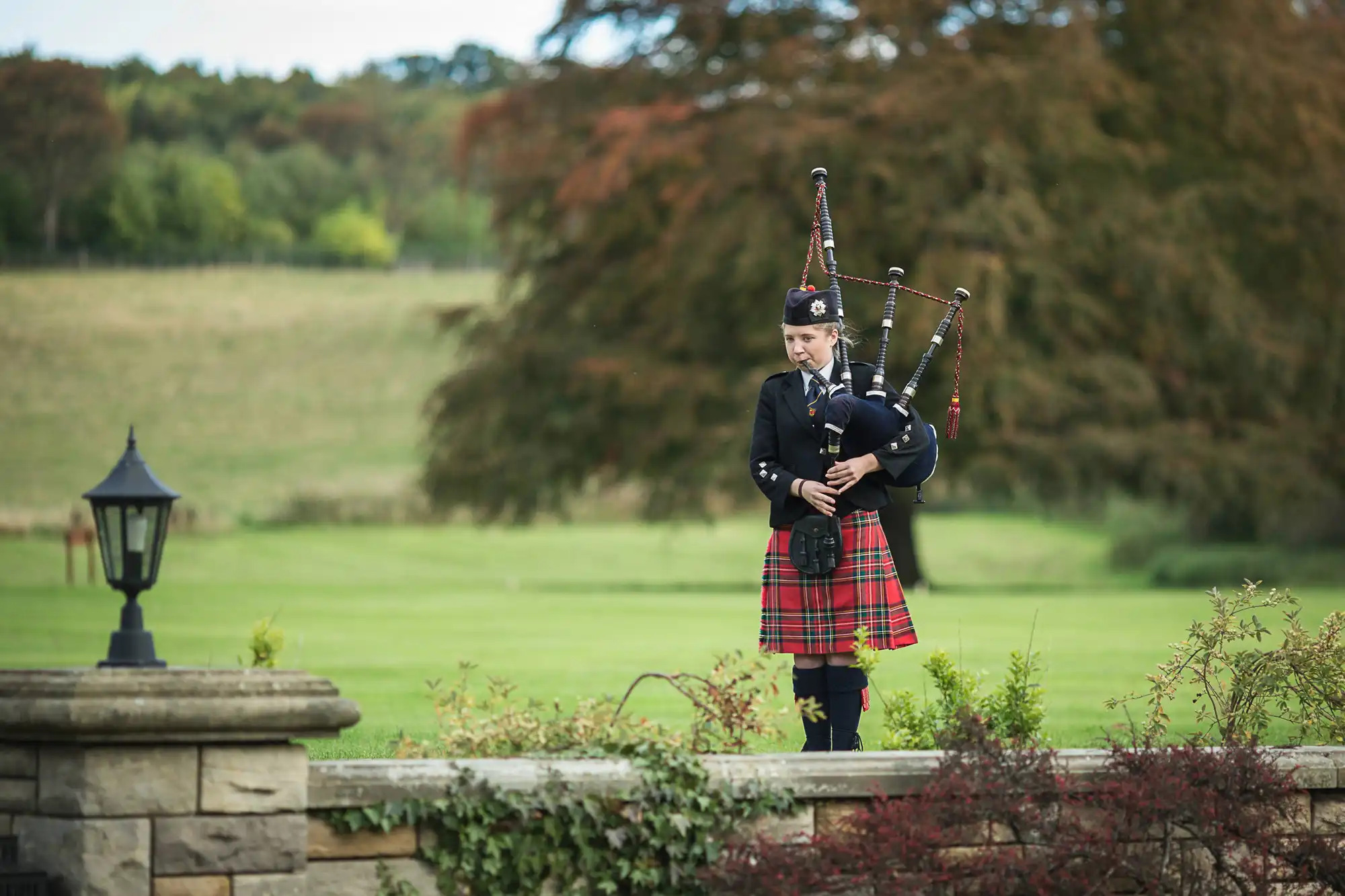 A bagpiper in traditional tartan kilt and uniform plays beside a stone wall with a lush, wooded backdrop.