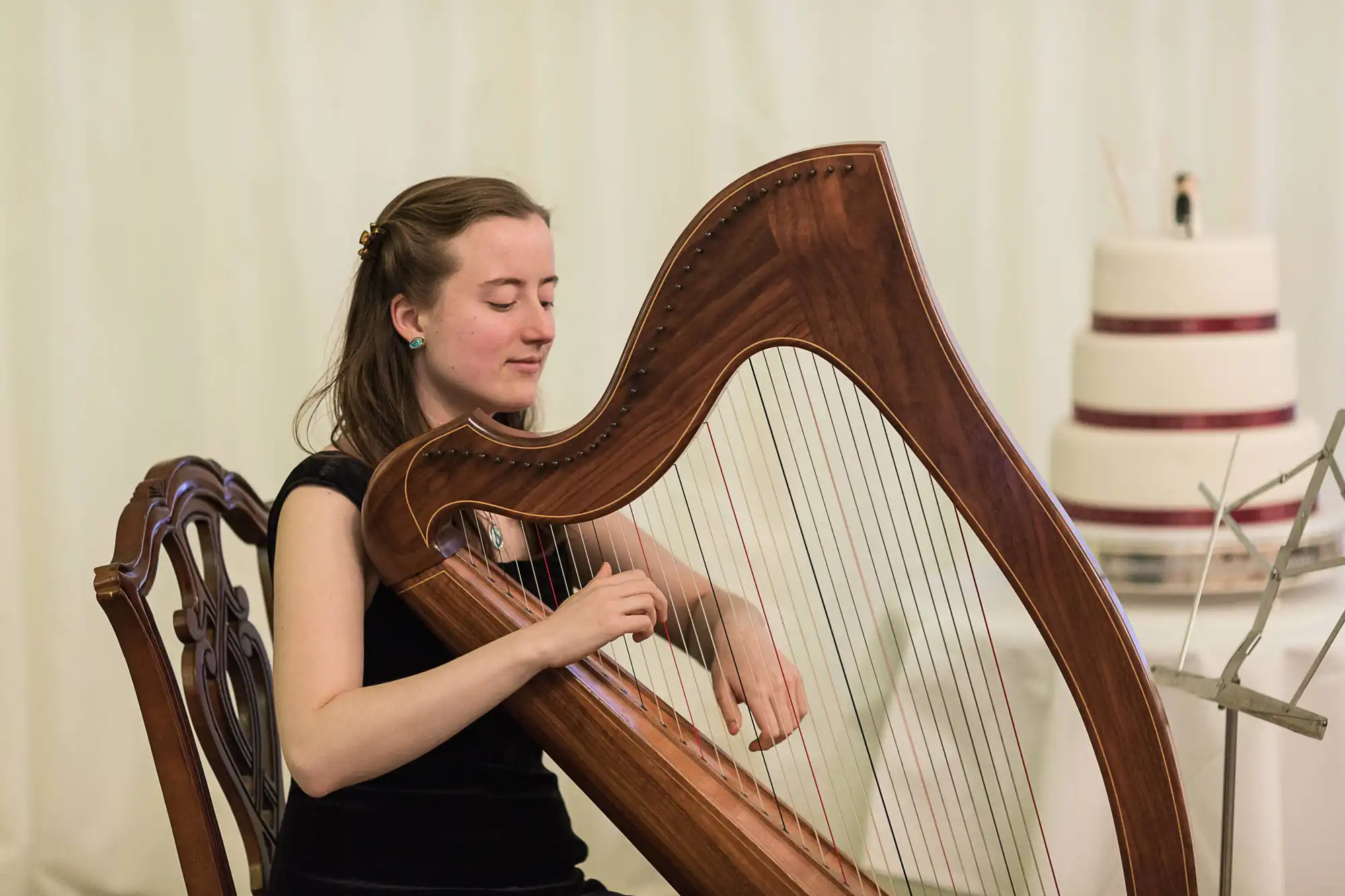 A woman plays a harp with concentration at a formal event, a tiered cake visible in the background.