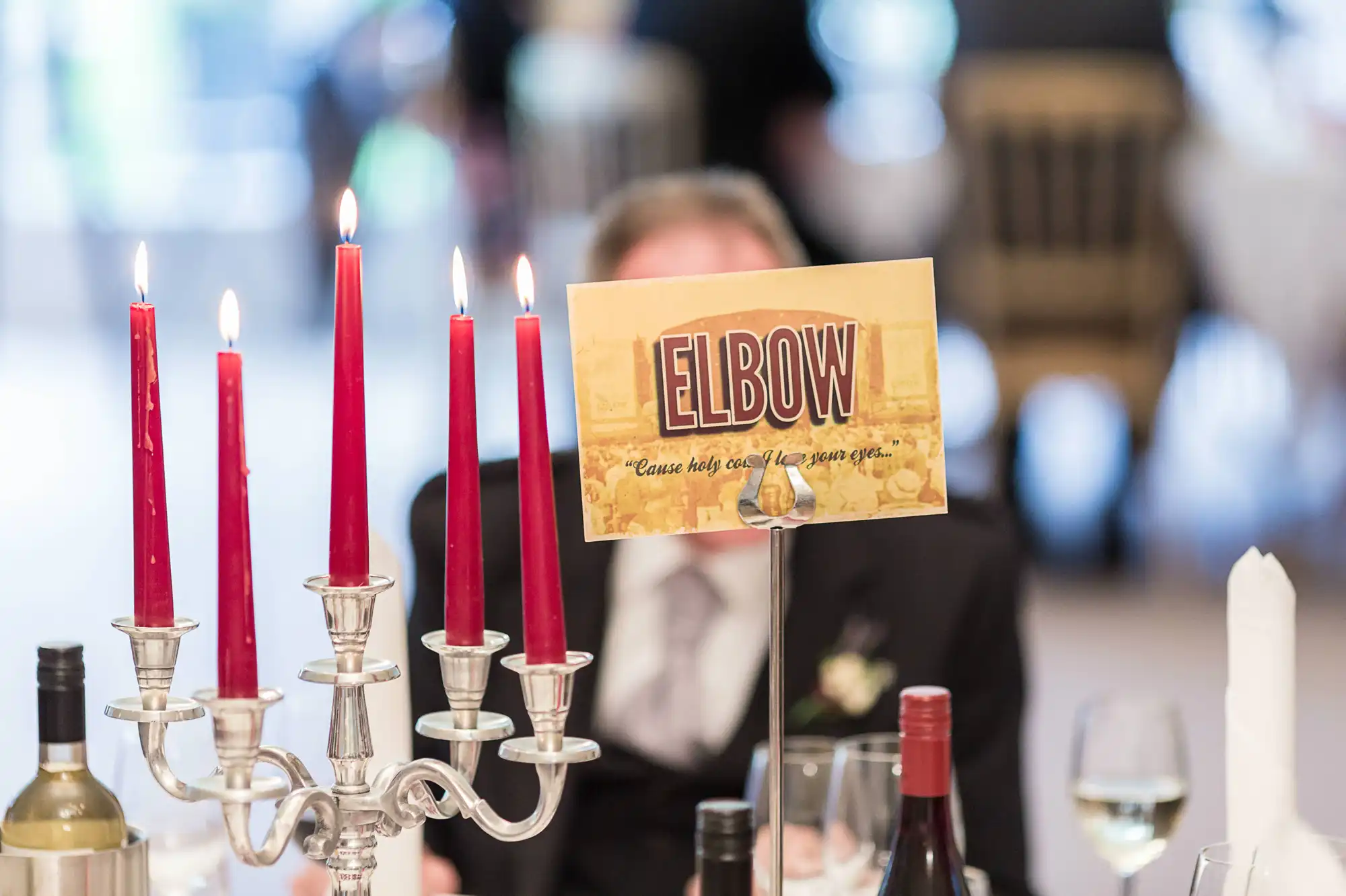 Elegant dining table setting with a silver candelabra holding red candles, a table sign reading "elbow," and blurred guest in the background.