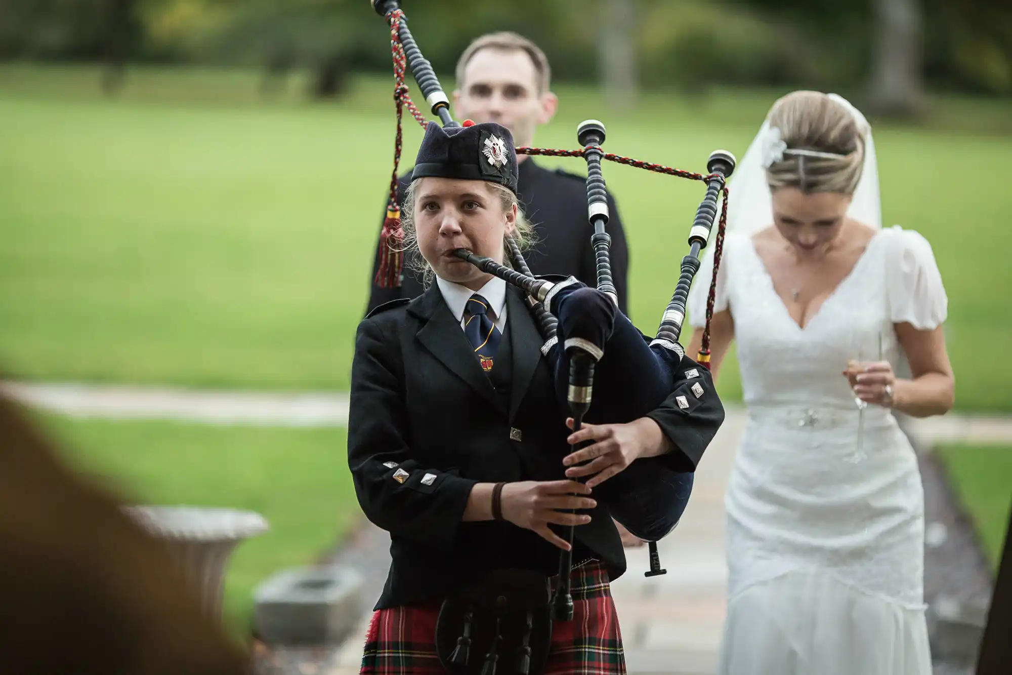 A young bagpiper in traditional scottish attire plays at a wedding, with a bride walking in the background.