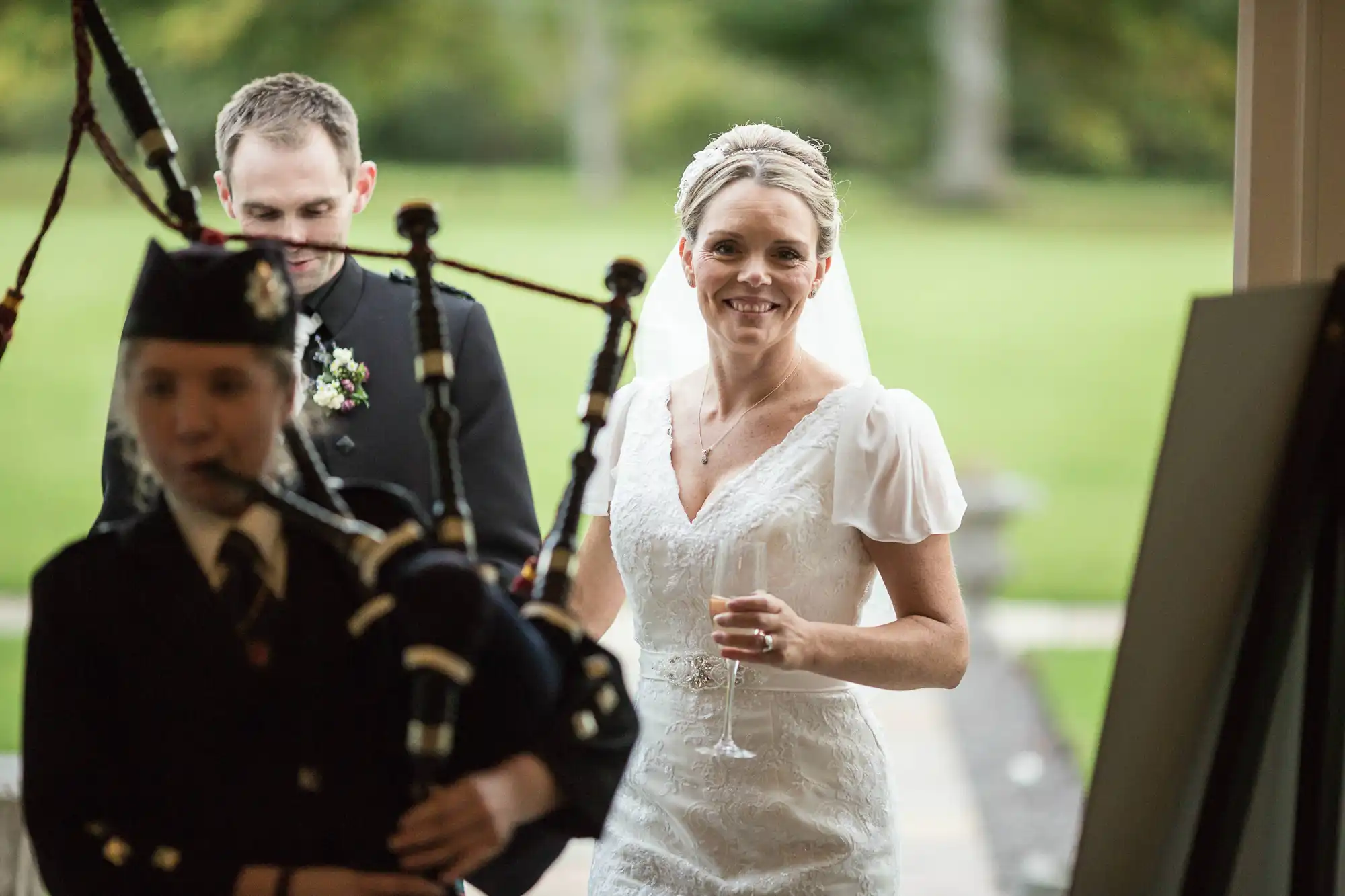 A bride holding a glass smiles as she walks past a bagpiper, with the groom standing in the background.