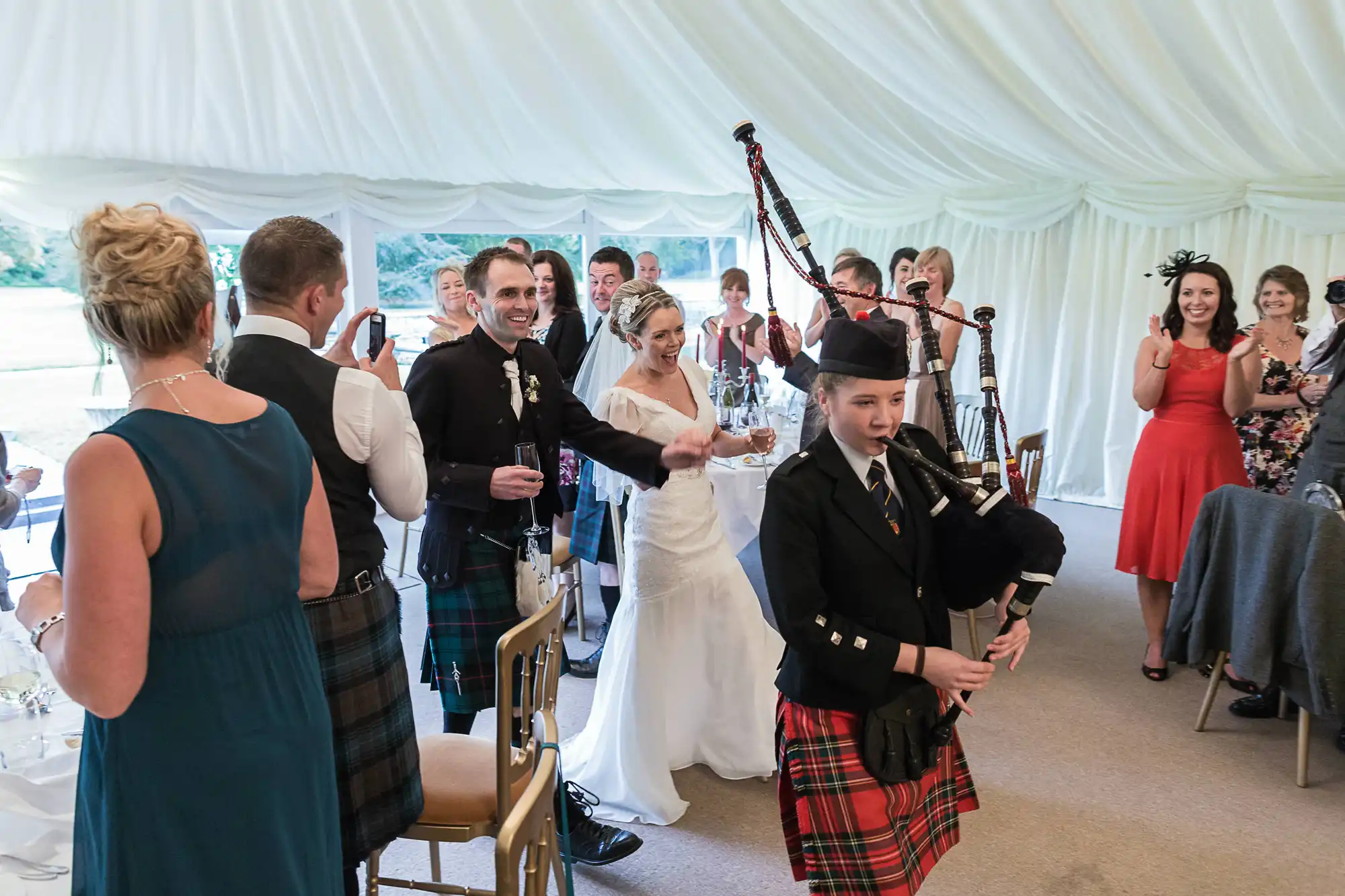 Wedding guests applauding as a bagpiper leads the newlyweds into the reception tent.