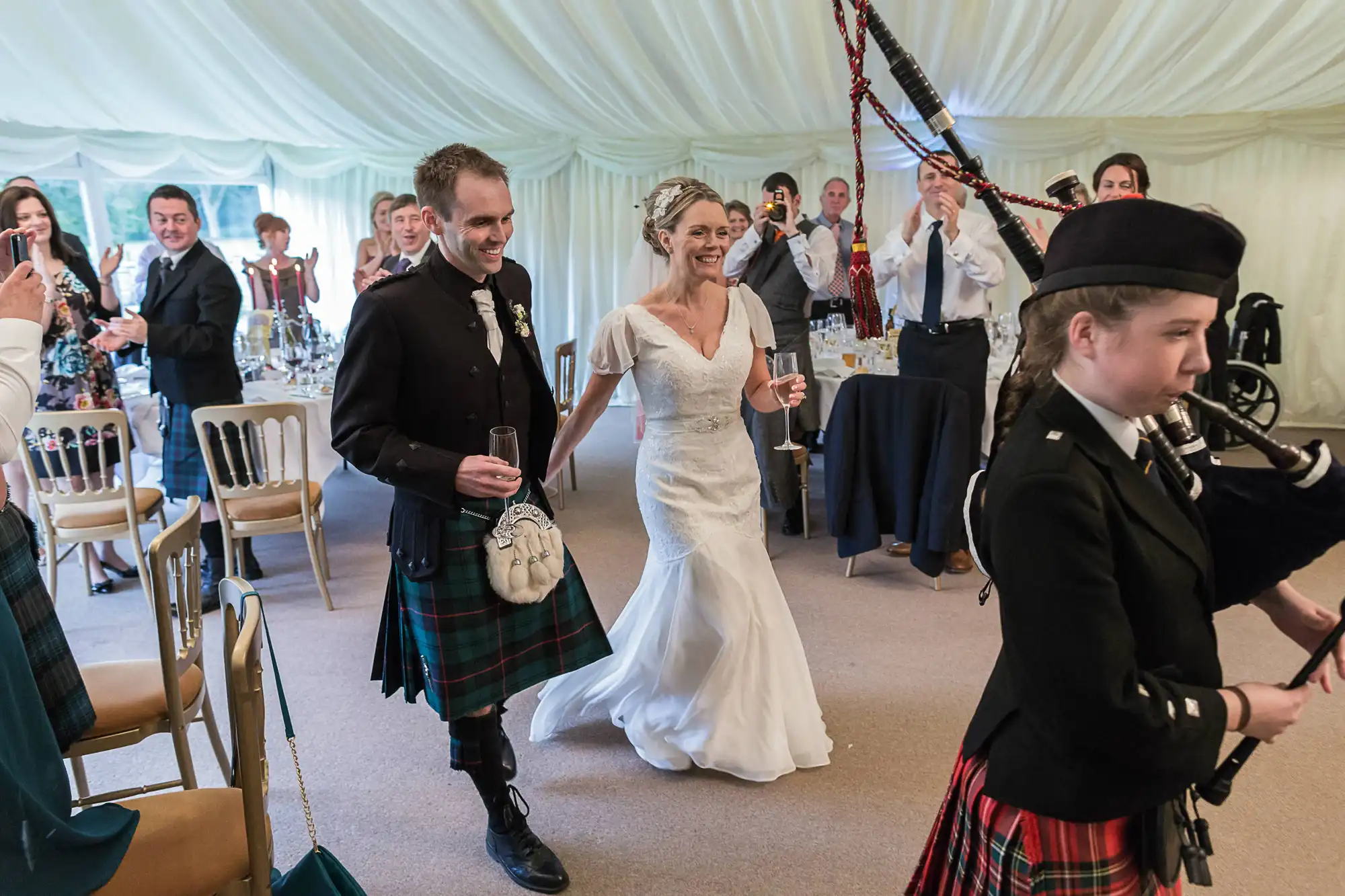 A bride and groom, both smiling, run through a wedding tent followed by a young bagpiper, as guests watch and clap.