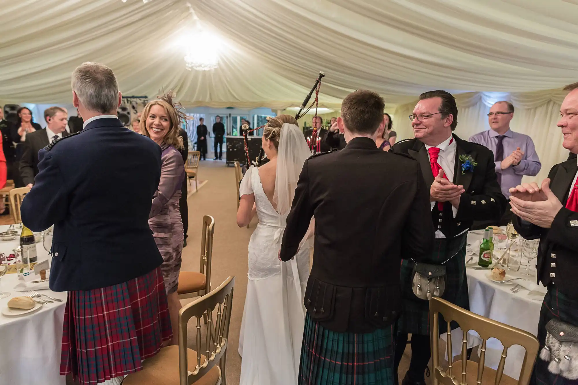 Guests in kilts applauding a bride and groom at a wedding reception inside a tent.
