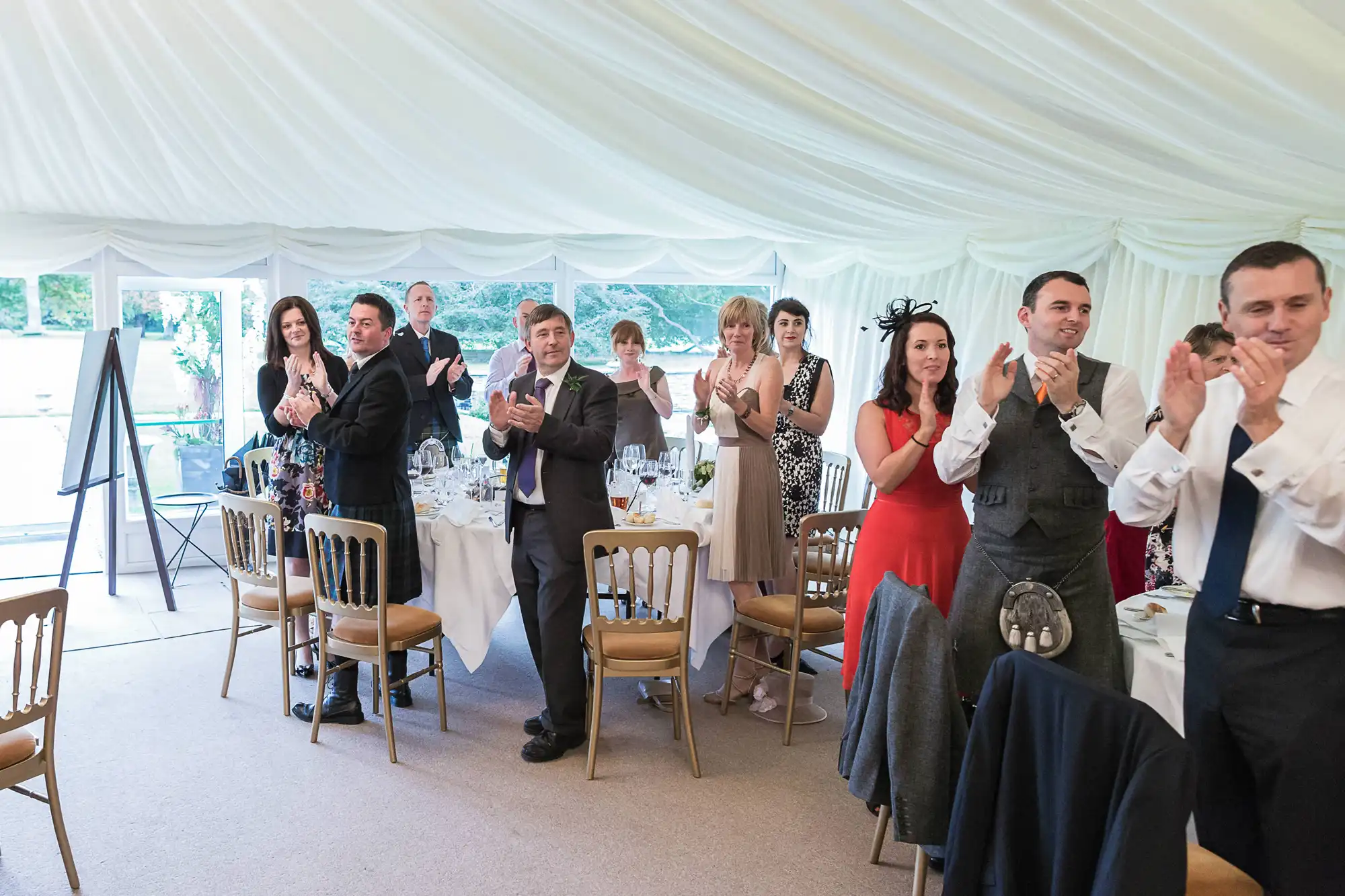 Guests standing and applauding at a wedding reception inside a tent.