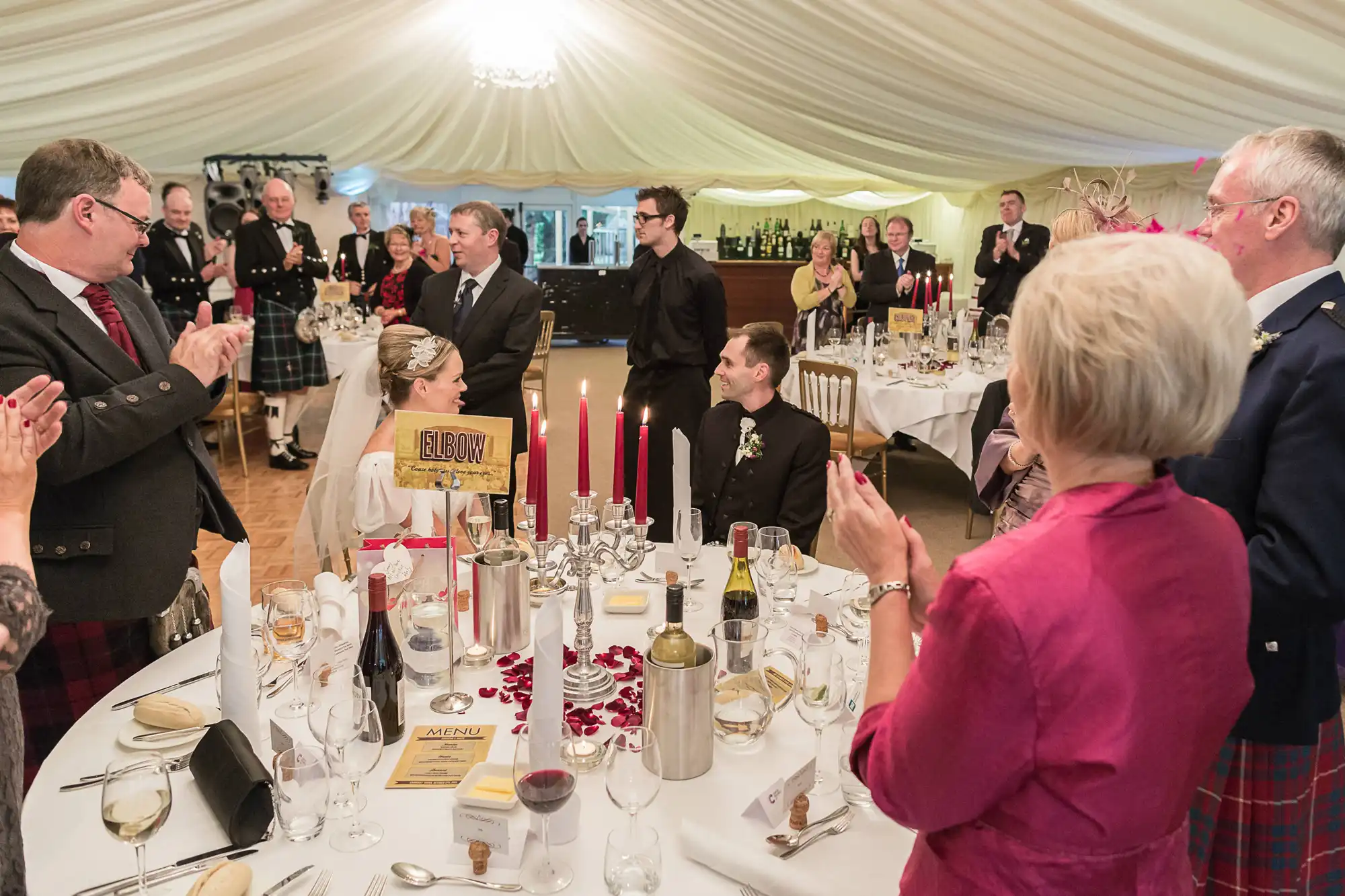Guests in formal attire, including kilts, clapping hands at a candlelit banquet table during an elegant event in a large tent.