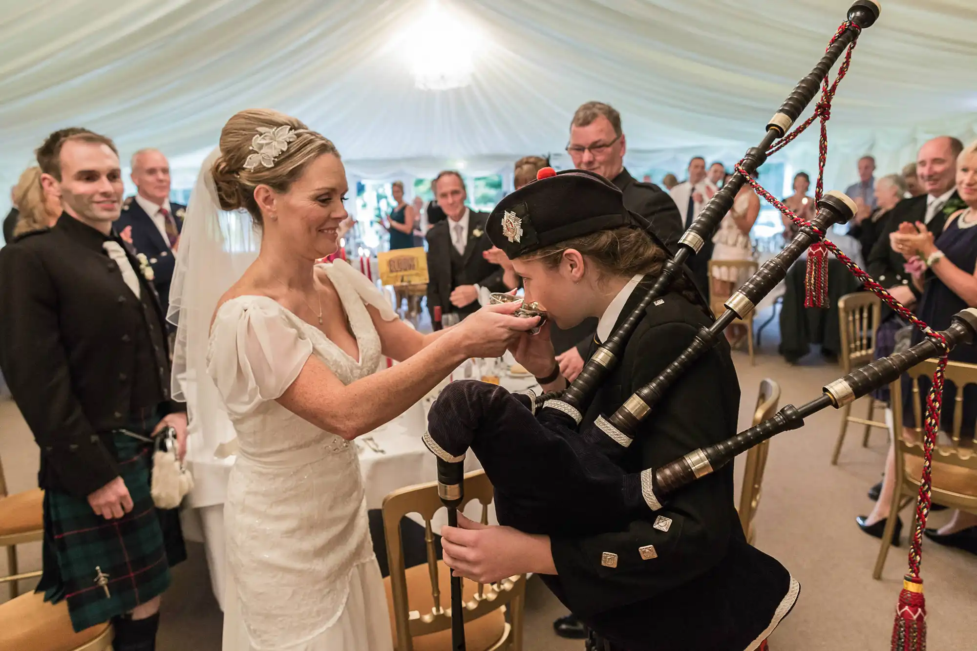 A bride in a white dress offers a drink to a young girl playing bagpipes at a wedding reception, with guests in formal attire watching.
