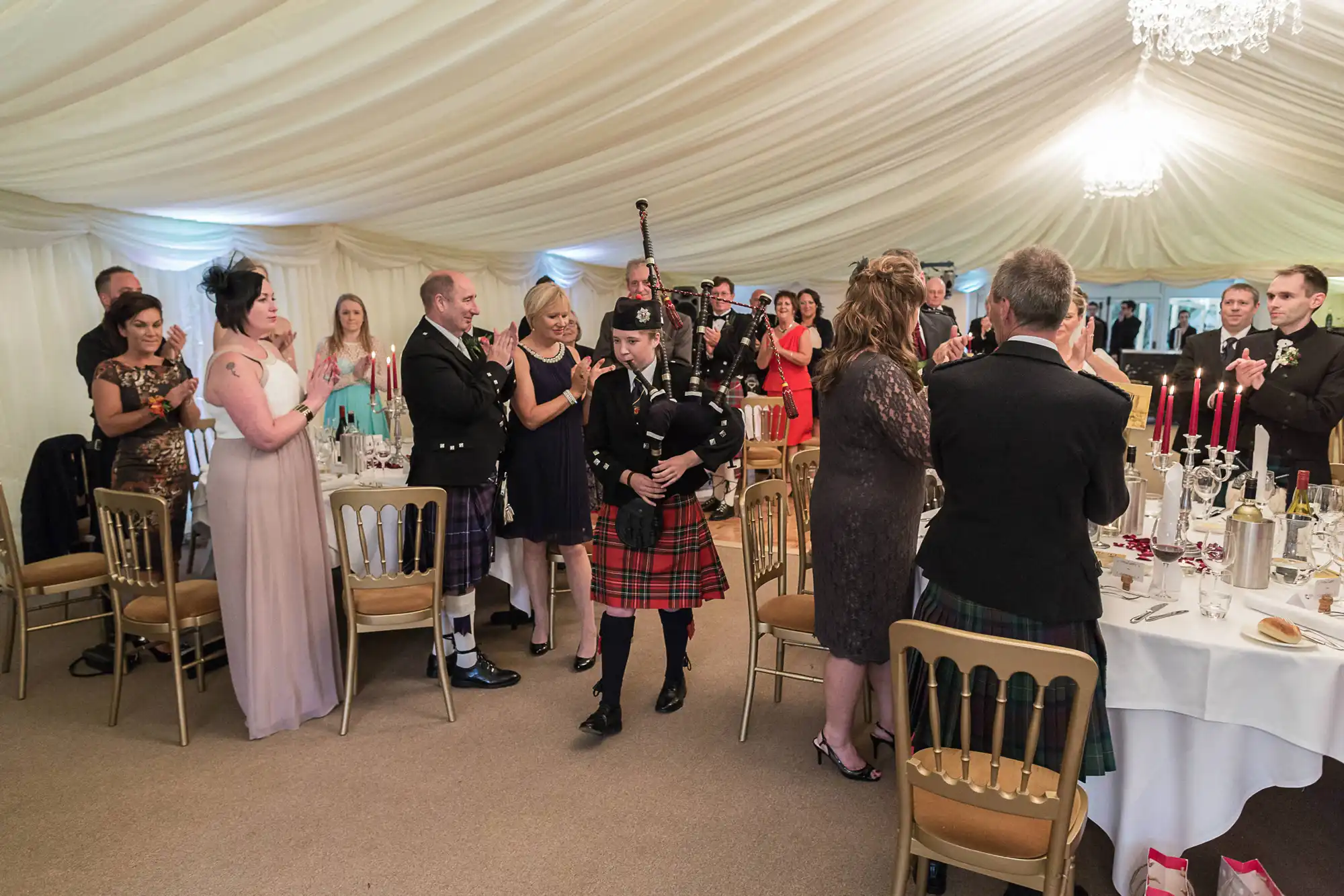 Guests applauding a bagpiper in traditional scottish attire walking through a banquet tent at a formal event.