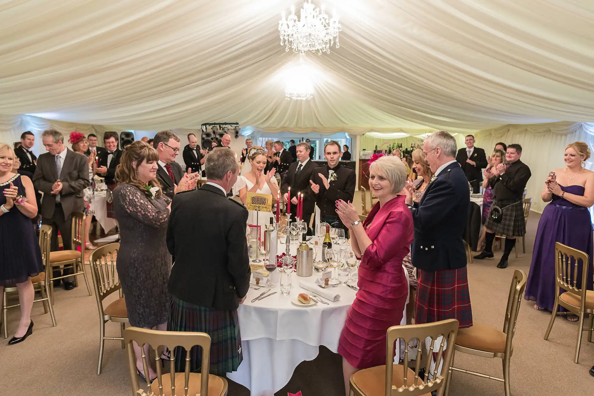 Guests in formal and semi-formal attire, including kilts, applauding at a wedding reception inside a tent with chandeliers.