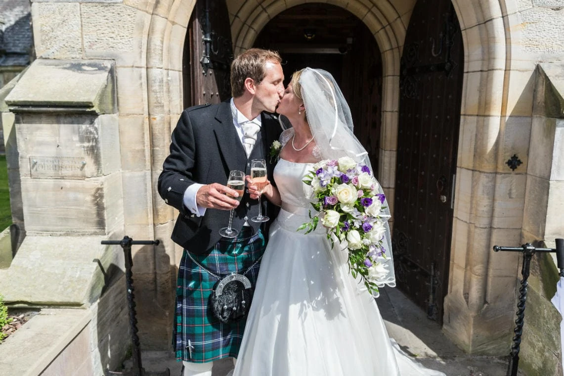 newlyweds kissing with a glass of champagne at the entrance to Carberry Tower