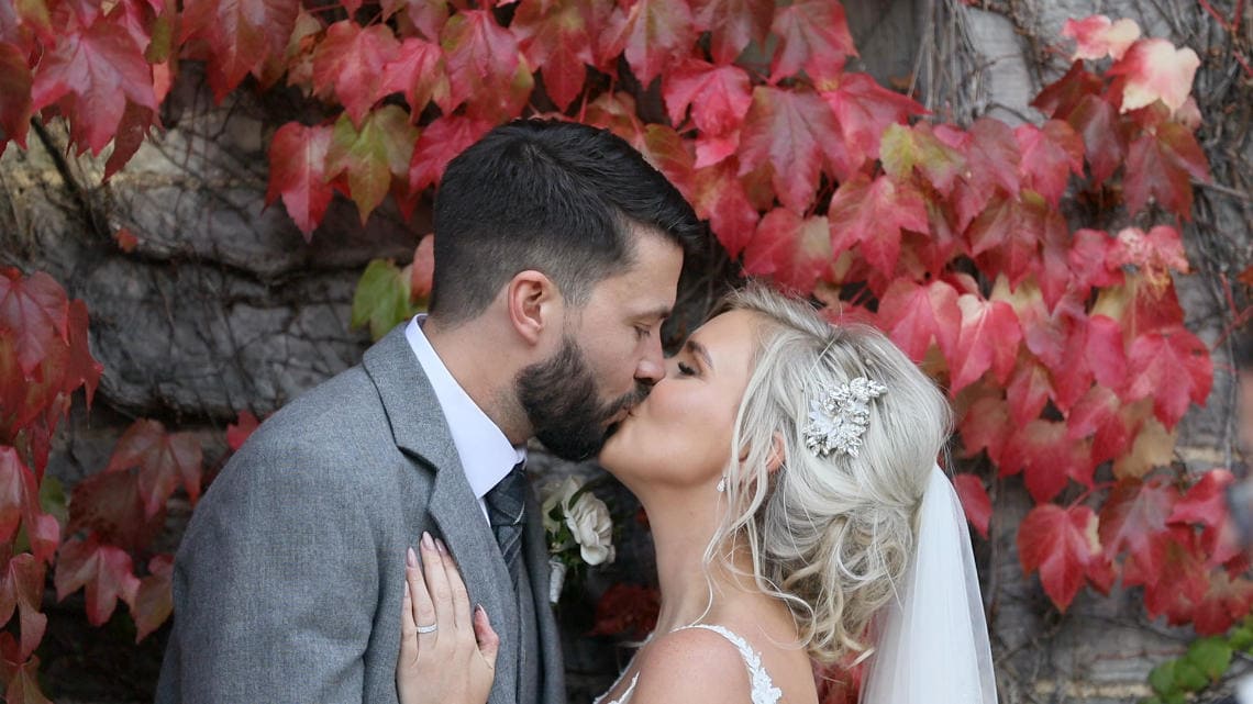 A bride and groom kiss at the altar in a rustic stone venue, surrounded by seated guests. An audience member claps, and a dog lays near the front on the right side.