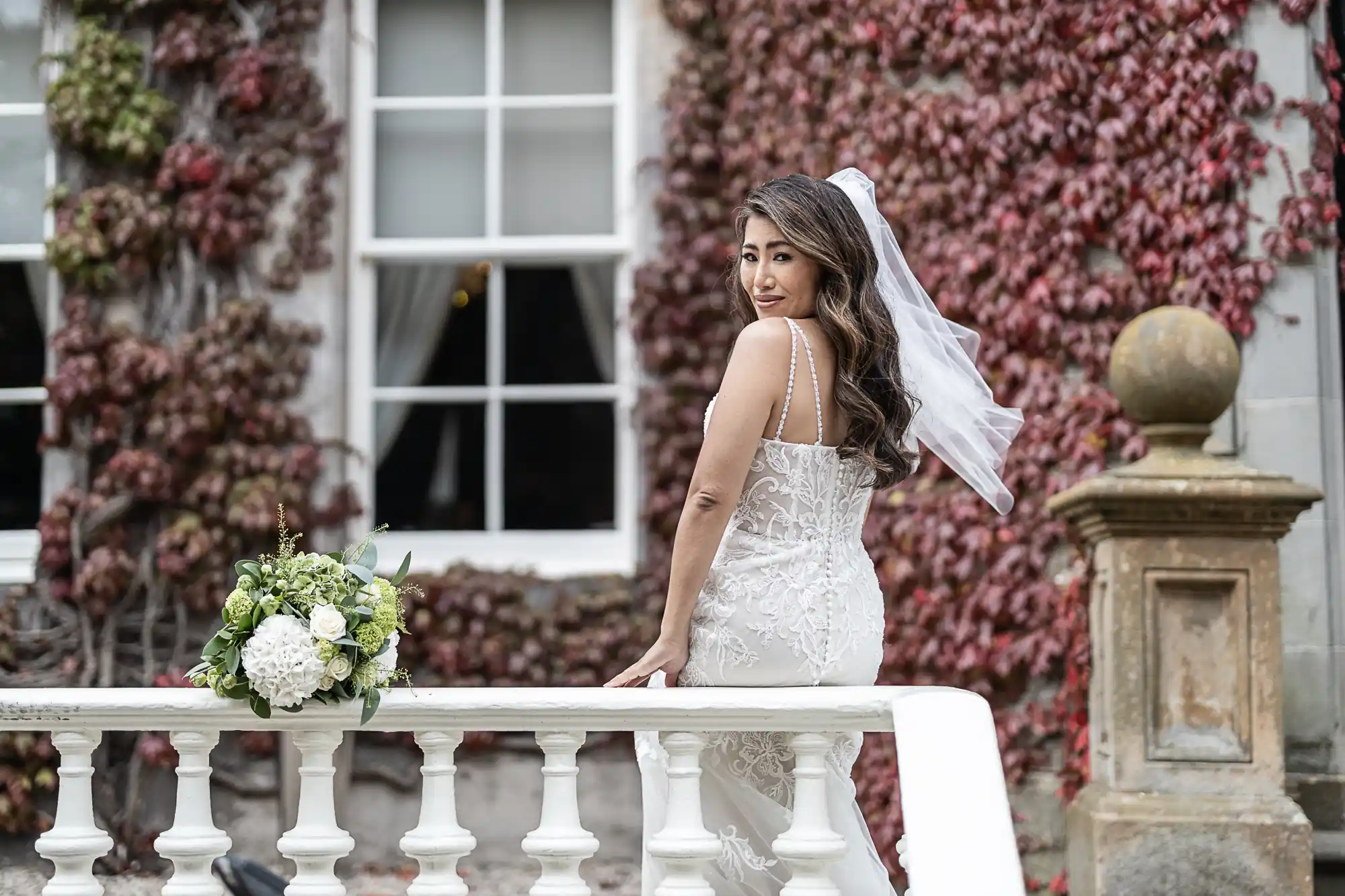 A bride in a white lace dress and veil stands by a white railing, with a bouquet of white and green flowers nearby. Red ivy covers the wall behind her, next to a large window.