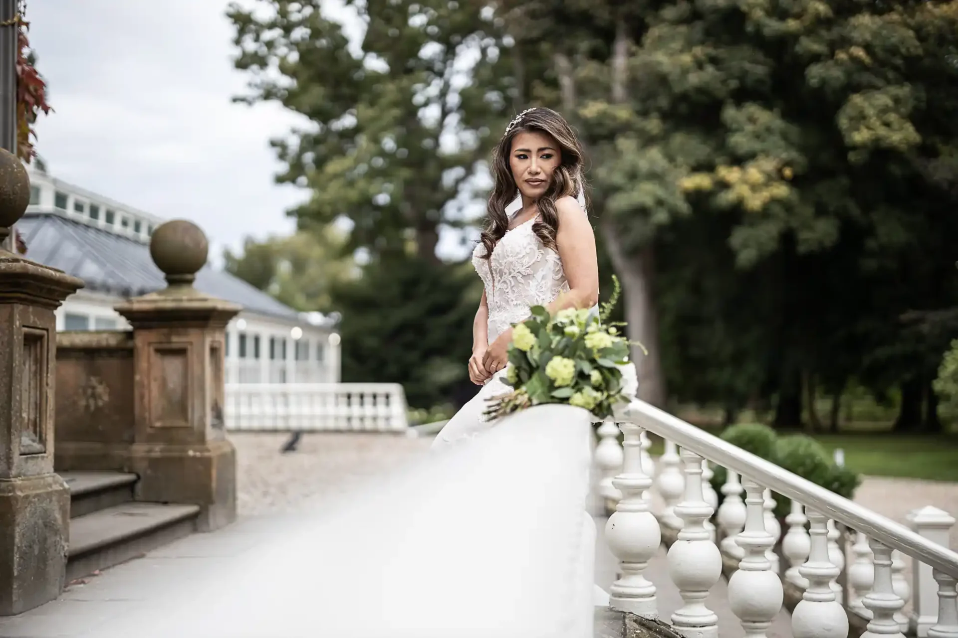 Bride in a white dress stands on an outdoor staircase beside a bouquet, with trees and a building in the background.